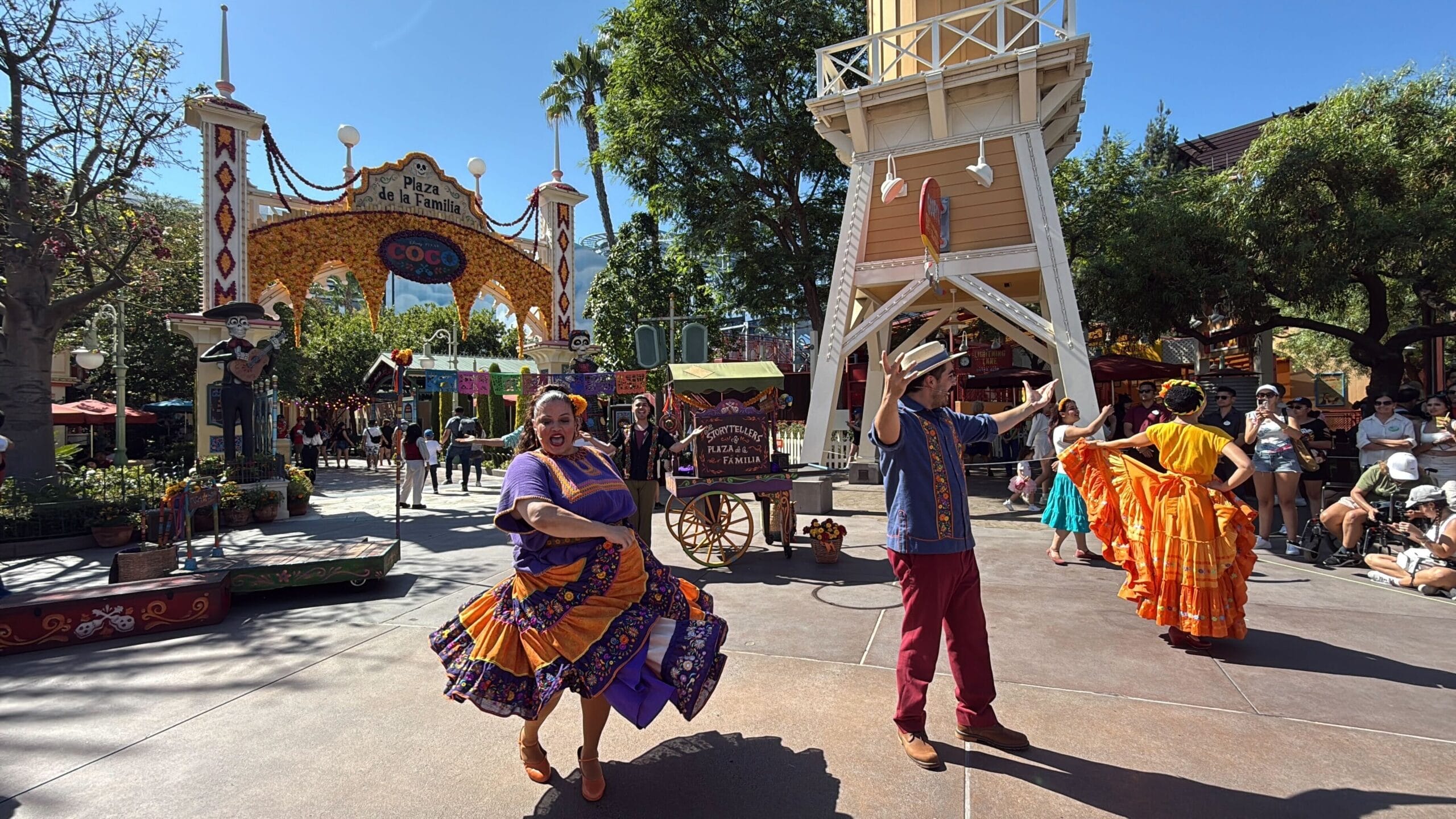 Dancers in colorful traditional outfits perform at Plaza de la Familia with Coco decorations and onlookers nearby.