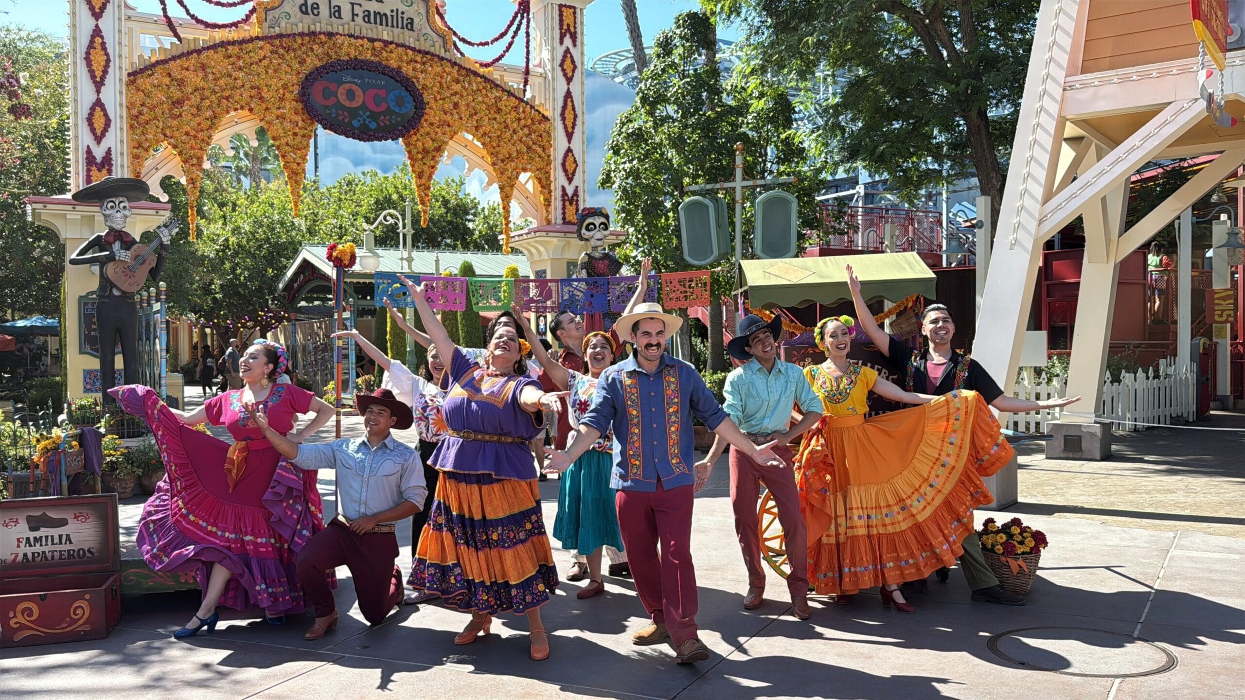 A group of performers in colorful costumes dance before a Coco-themed arch at Plaza de la Familia in Disney California Adventure.