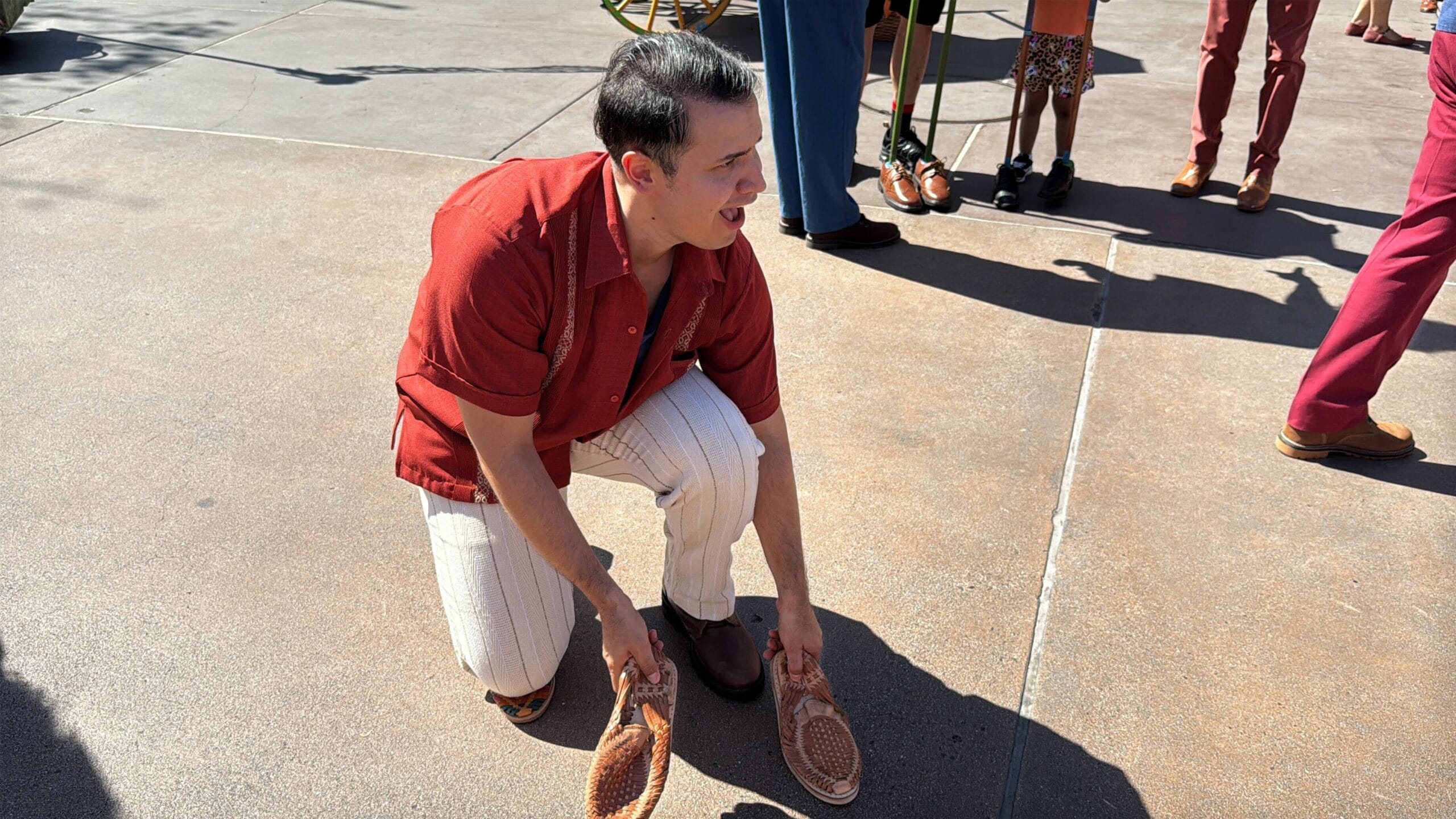 A person in a red shirt and striped pants kneels on the Plaza de la Familia sidewalk, holding woven sandals.