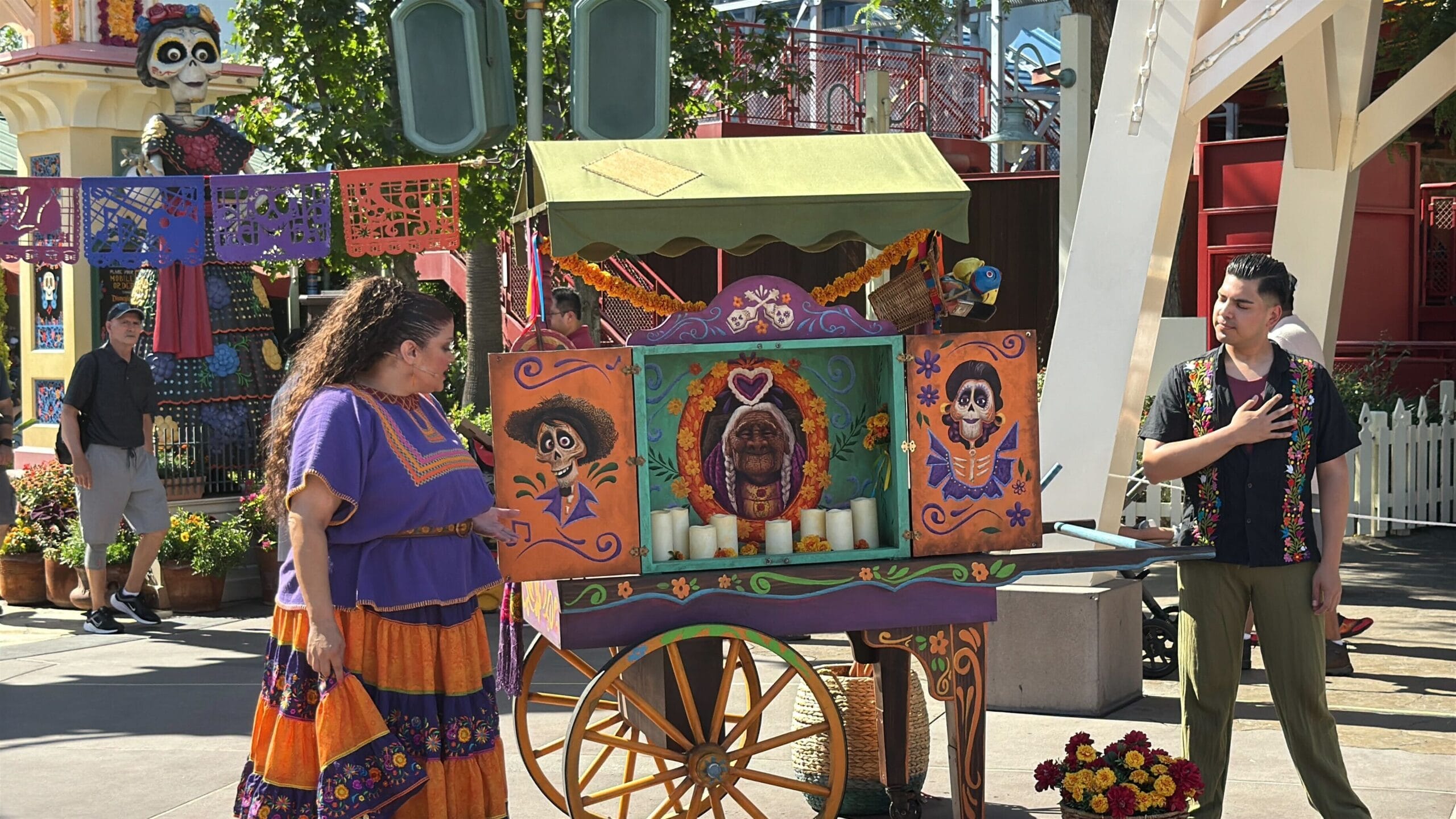 Two performers stand by a Día de los Muertos altar cart at Plaza de la Familia in Disney California Adventure.