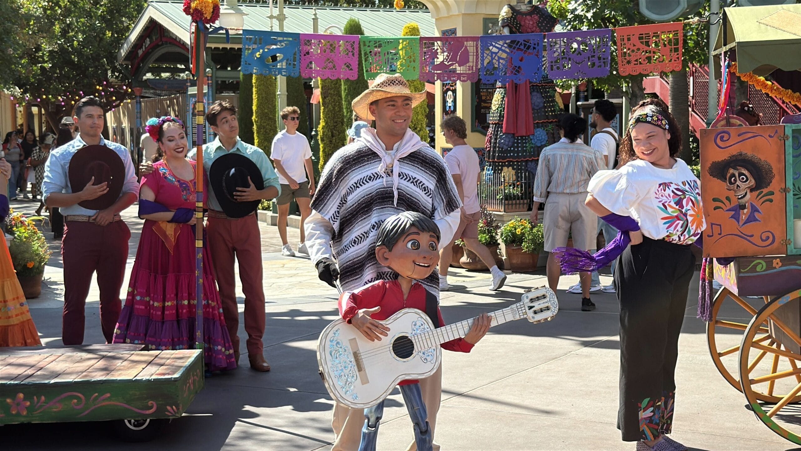 Performers in colorful costumes join Coco at Plaza de la Familia during a festive street show with papel picado decorations.