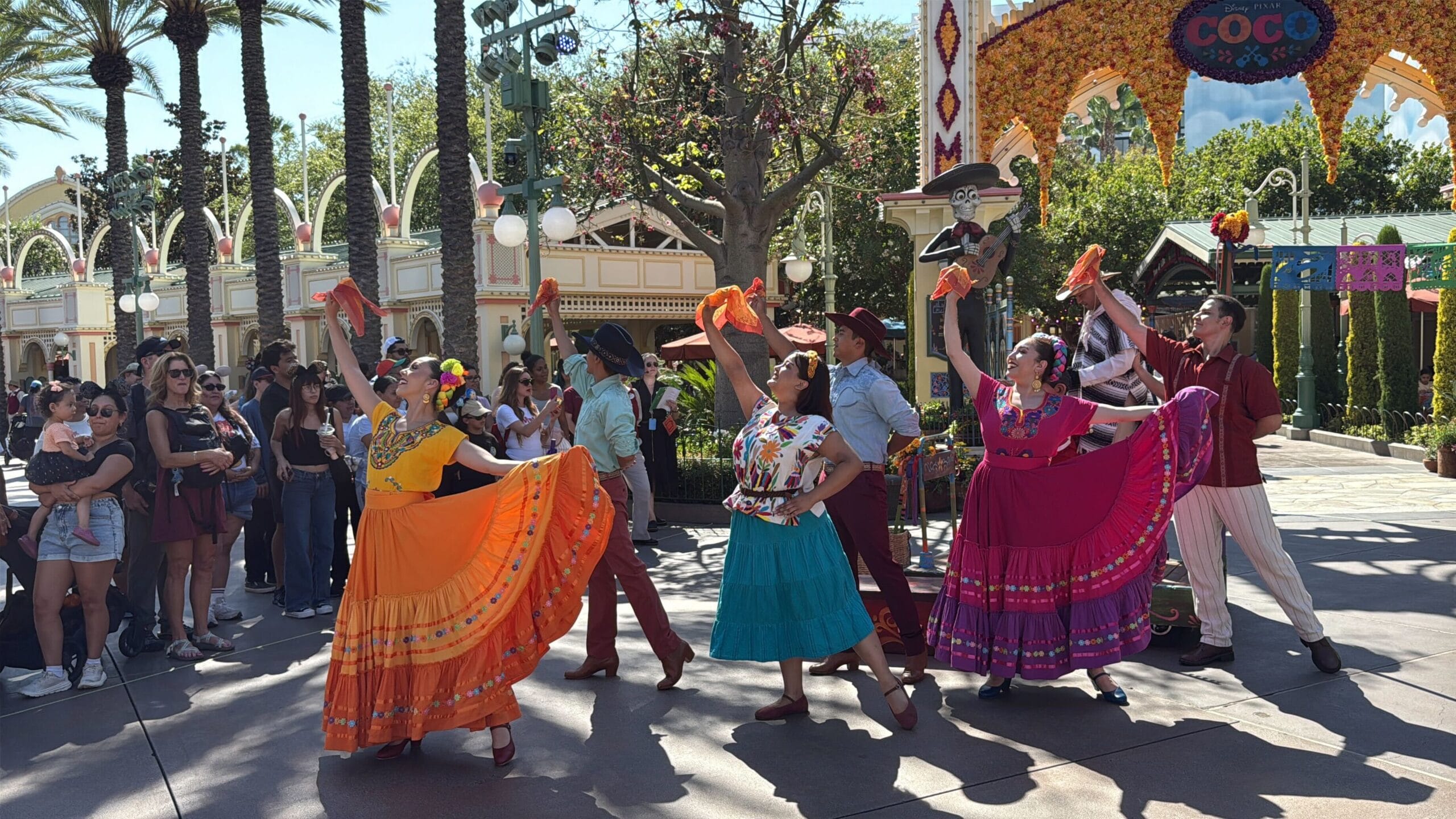 Performers in colorful traditional dresses dance with fans at Plaza de la Familia during a lively Coco celebration.