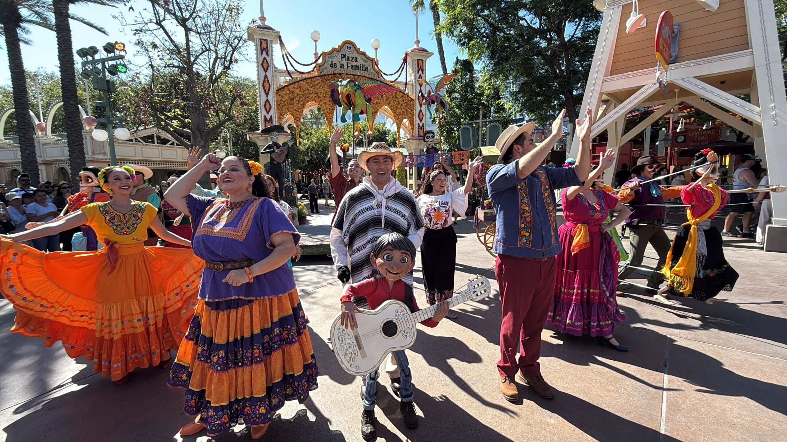Colorful parade at Plaza de la Familia with Coco dancers and a puppet boy holding a guitar, celebrating in festive streets.