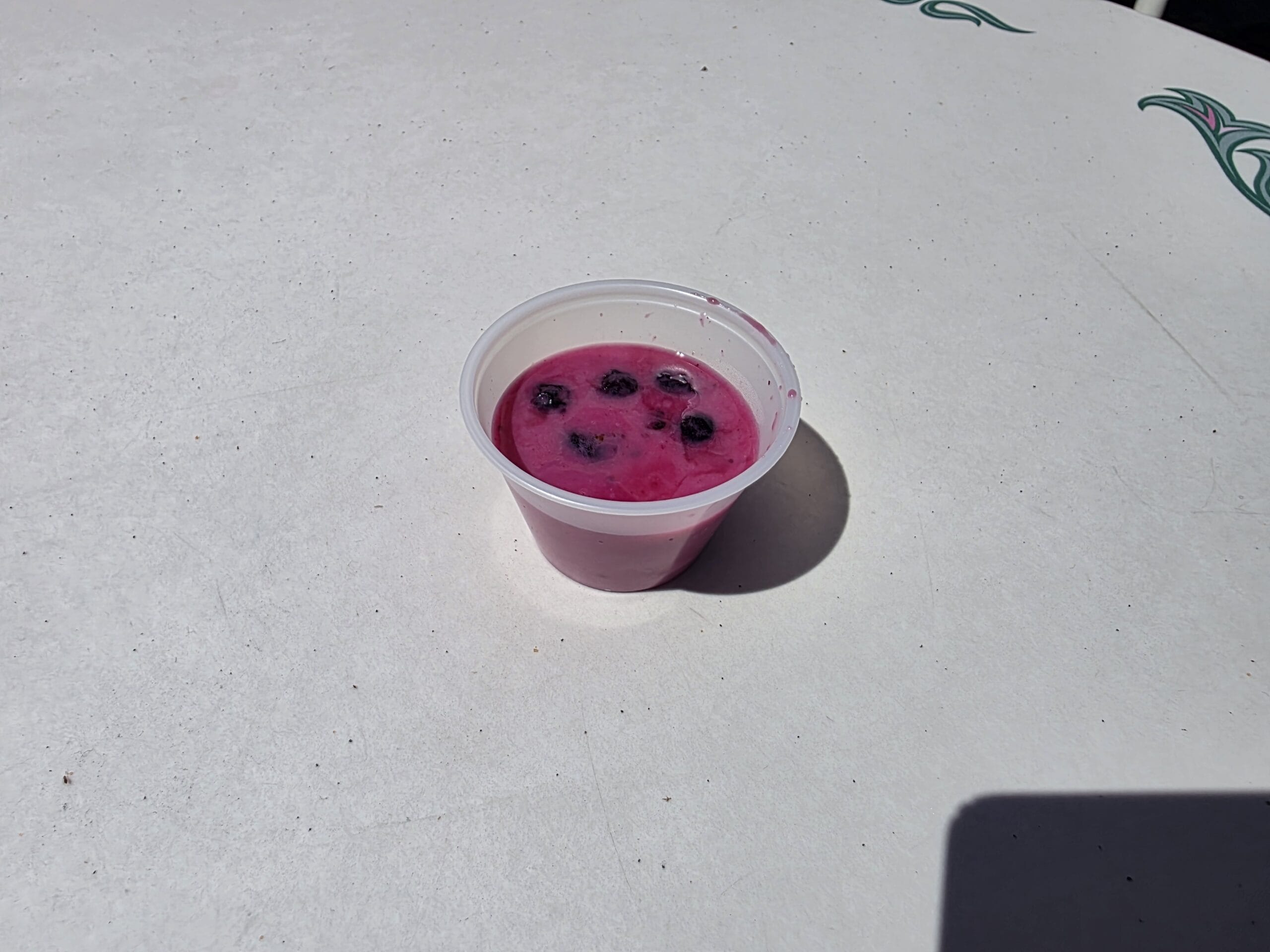 A small plastic cup with bright pink dipping sauce and dark berries sits on a white table in sunlight.