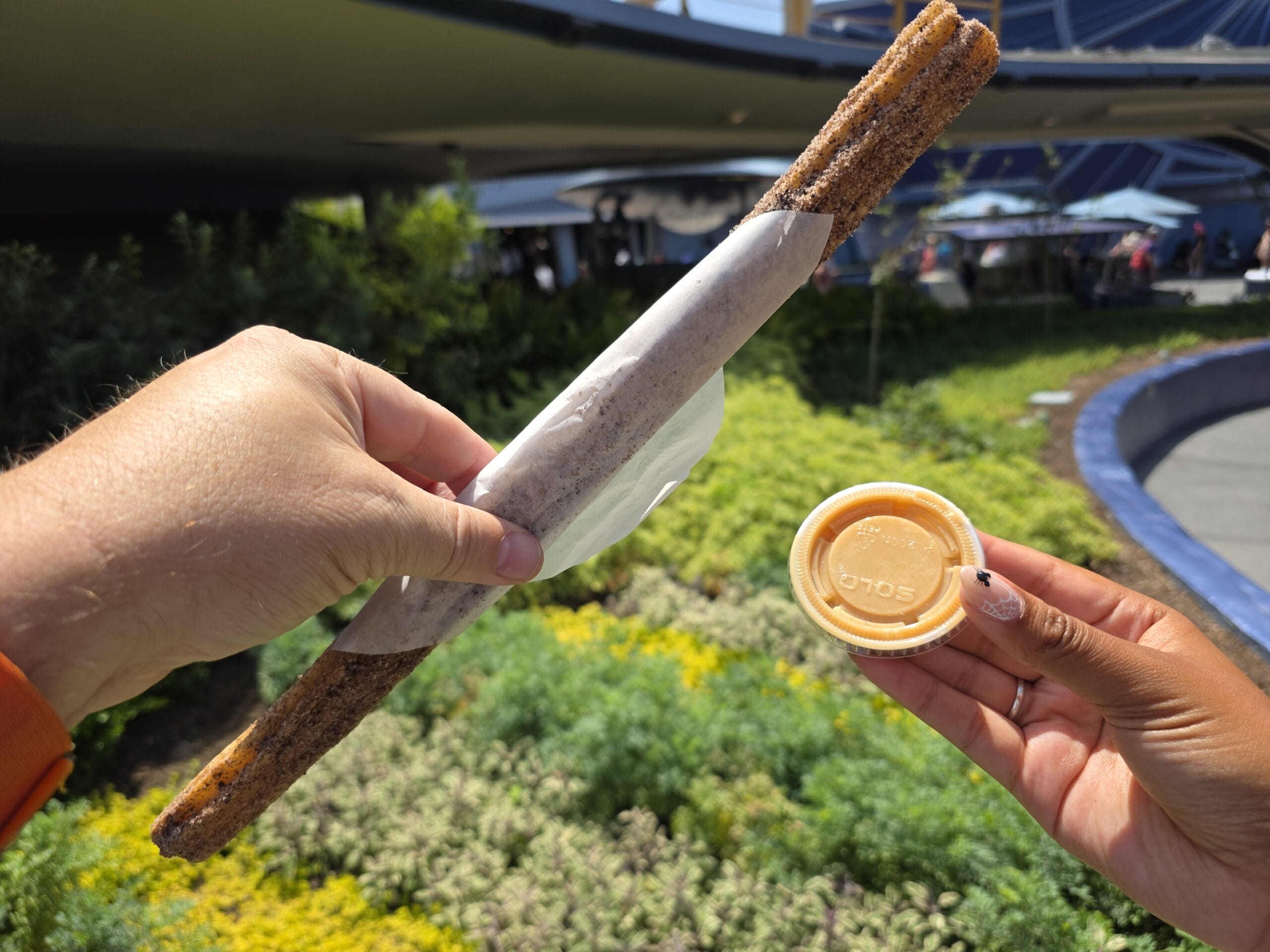 A hand holds a Halloween Churro at Disneyland, with another hand dipping it into festive orange dipping sauce outdoors.