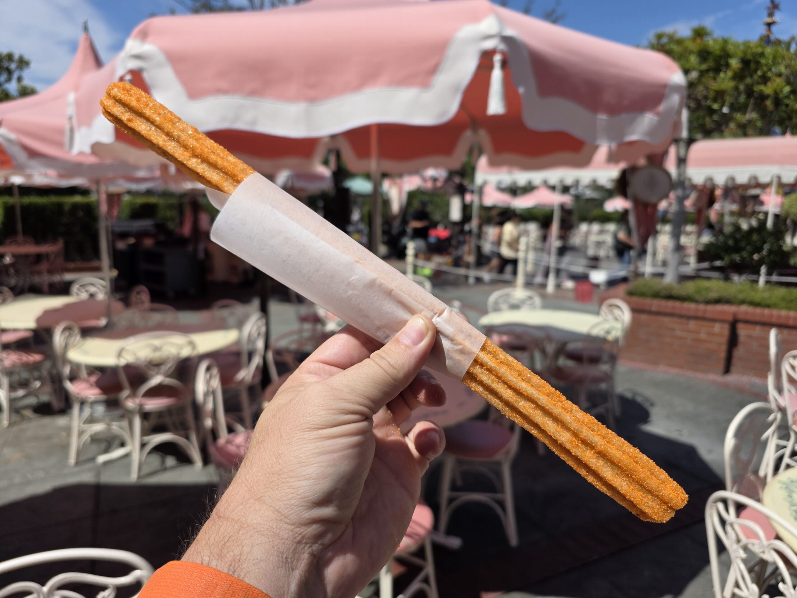 A hand holding a Halloween Churro at Disneyland, in front of outdoor tables with pink umbrellas and chairs.