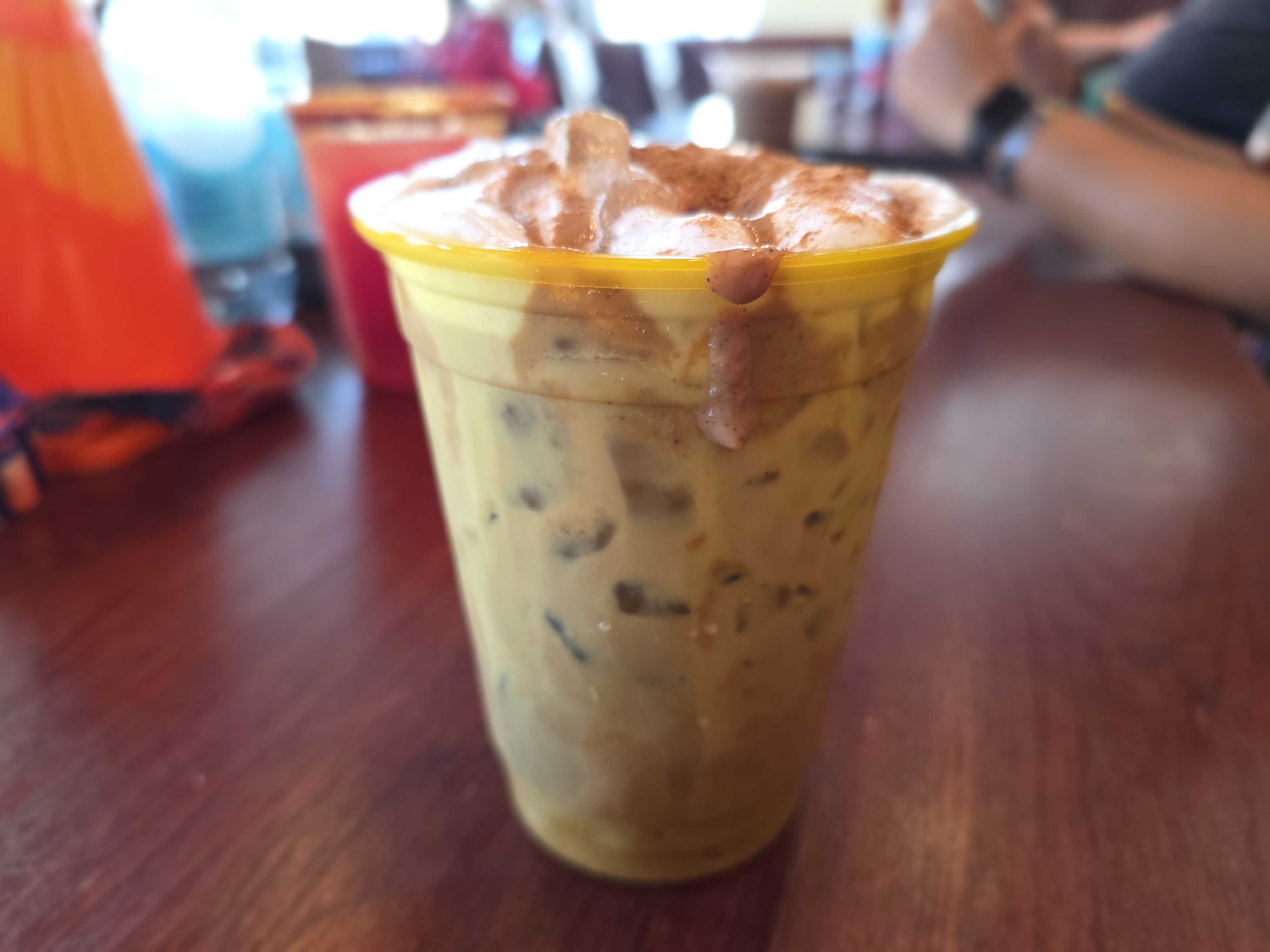 A plastic cup of iced coffee with foam sits on a wooden table at the Cozy Cone Motel café.