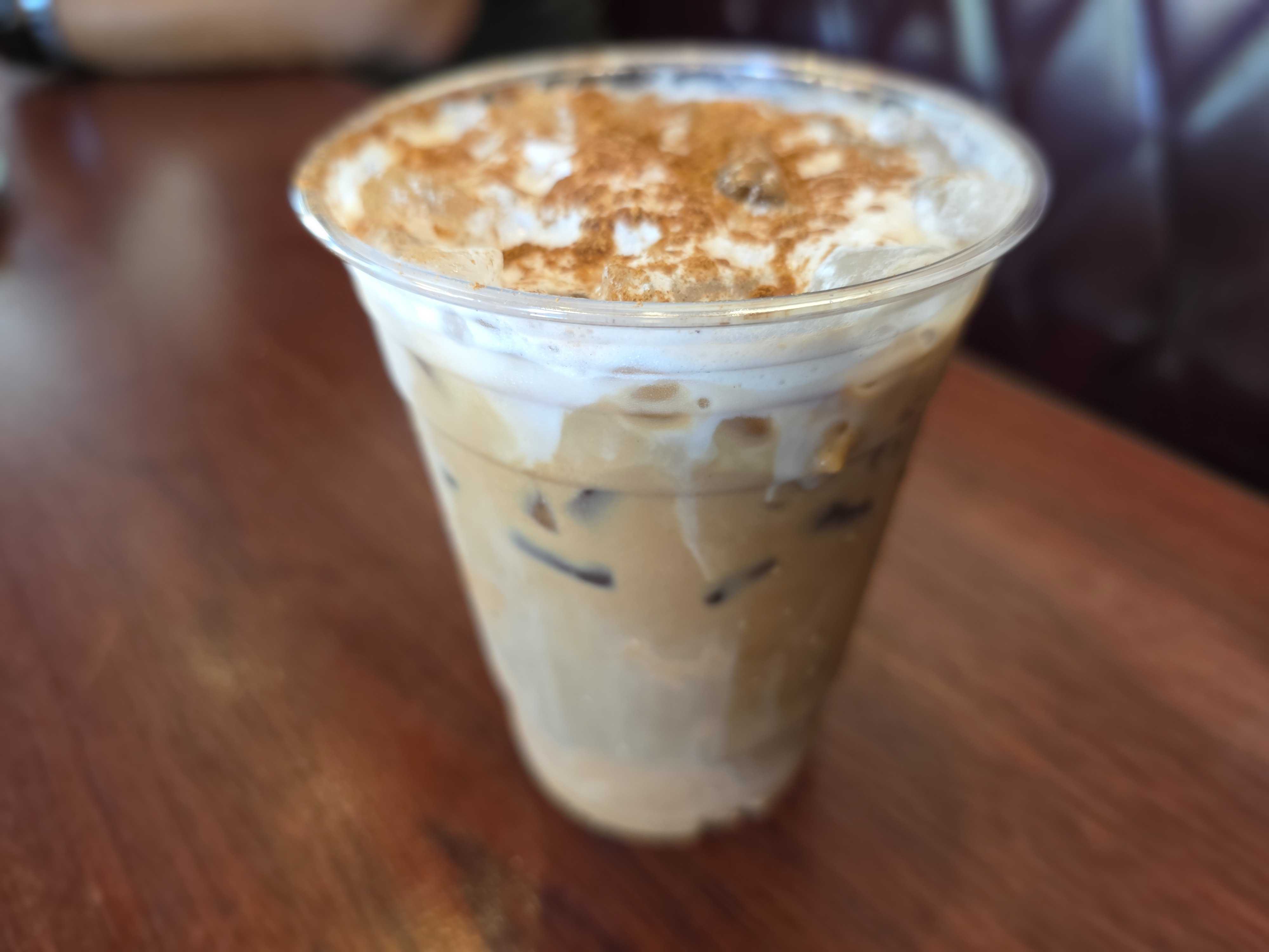 A Cozy Cone Motel plastic cup of iced coffee topped with whipped cream and cinnamon sits on a wooden table.