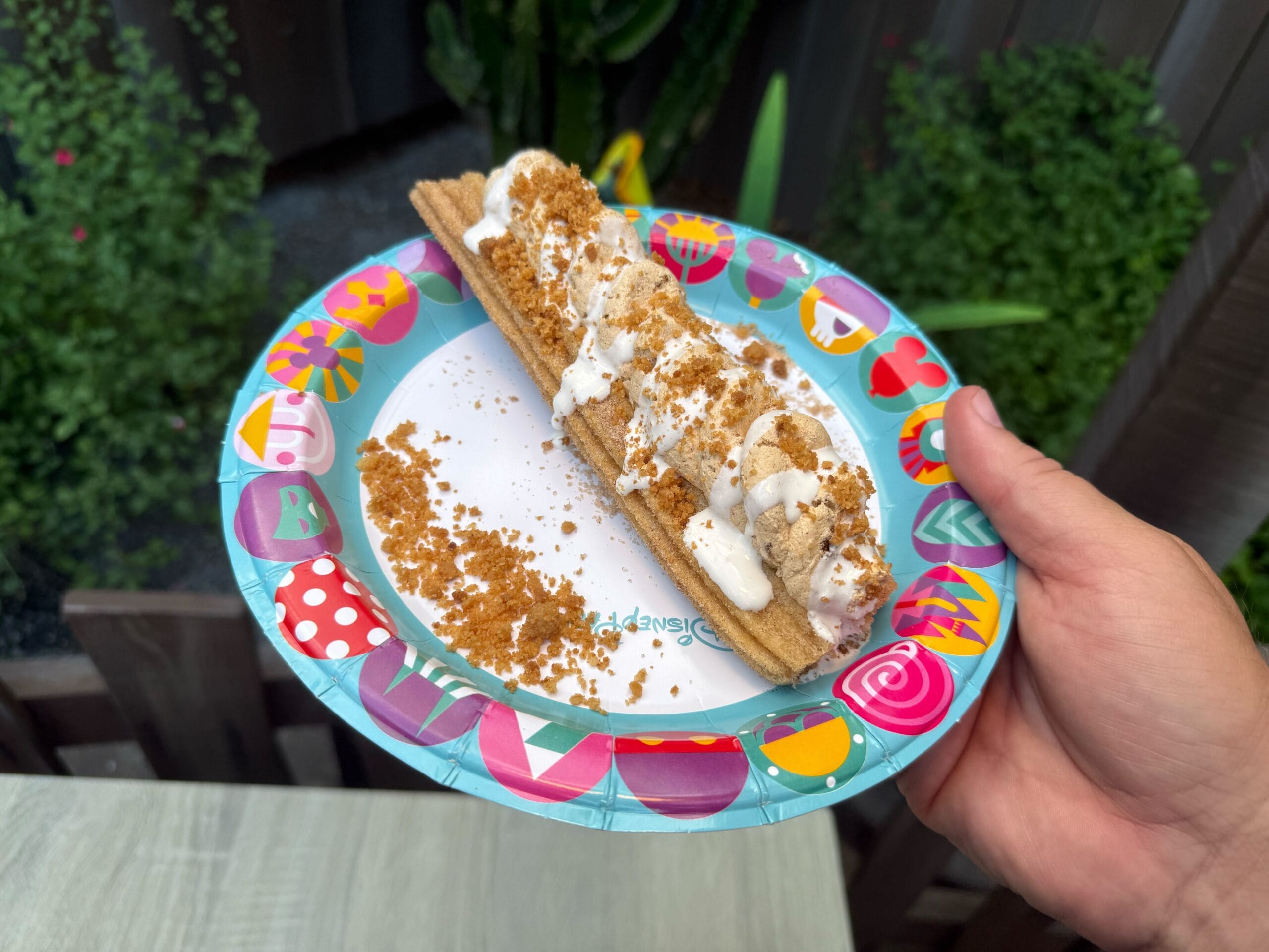 A hand holds a paper plate with a Cozy Cone Motel churro topped with ice cream and crumbled cookies, outdoors.