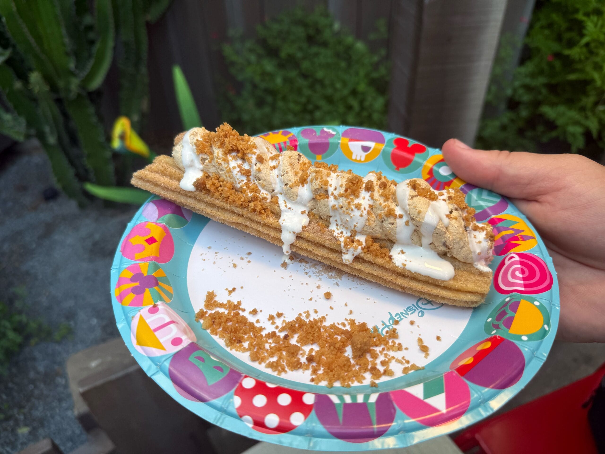 A Cozy Cone Motel churro with white icing and crumbs on a colorful plate, held outdoors near plants.