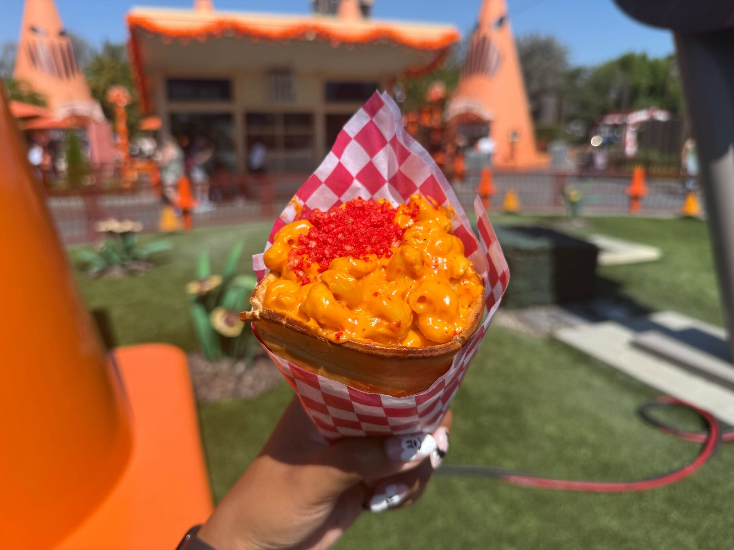 A hand holds a cone of mac and cheese with crunchy red bits outside the cozy, orange-themed Cozy Cone Motel.