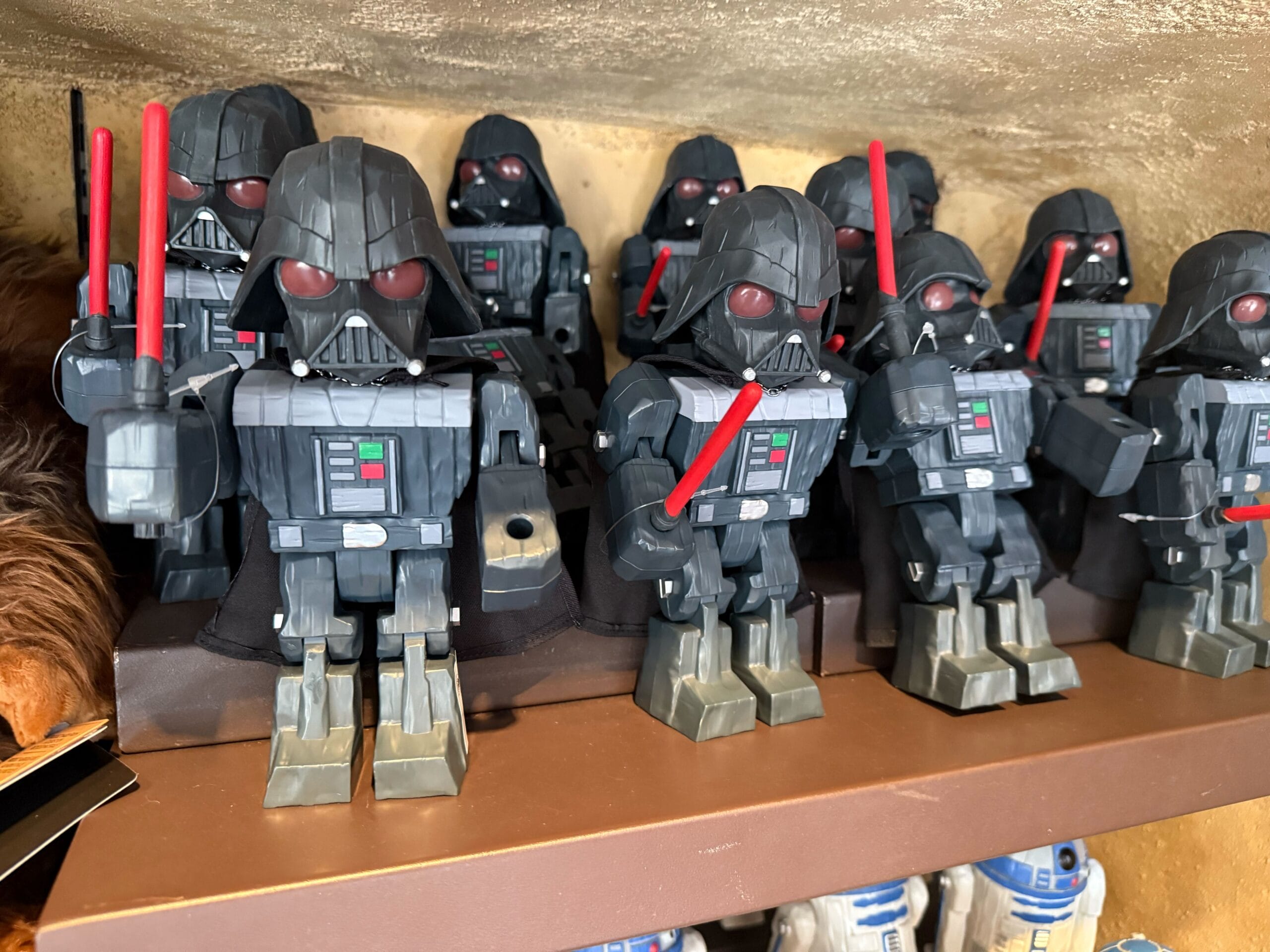 Several Darth Vader toy figures holding red lightsabers are lined up on a shelf with more toys in the background.