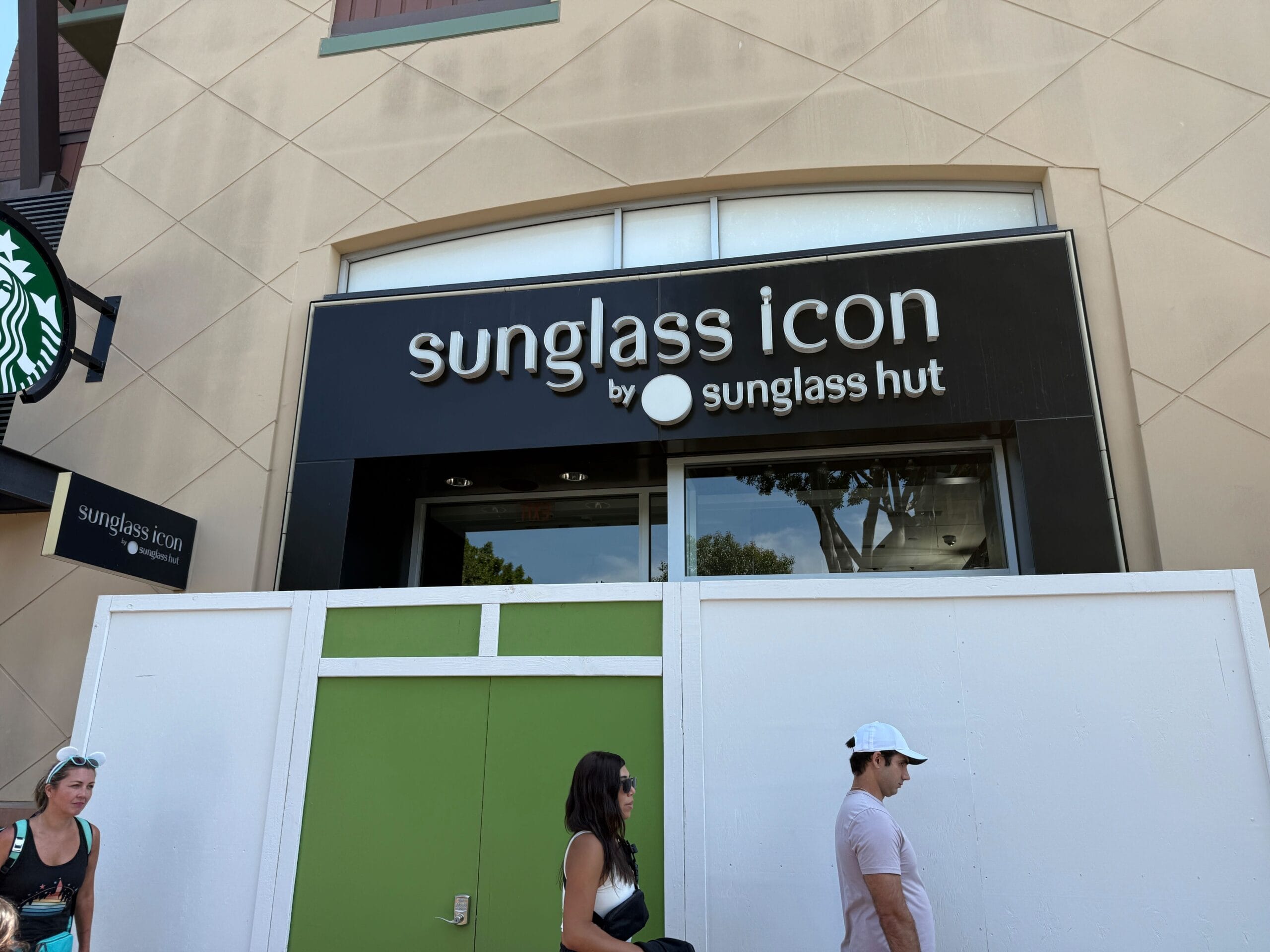 Storefront sign reads “sunglass icon by sunglass hut”; three people walk past a green and white barrier in front.