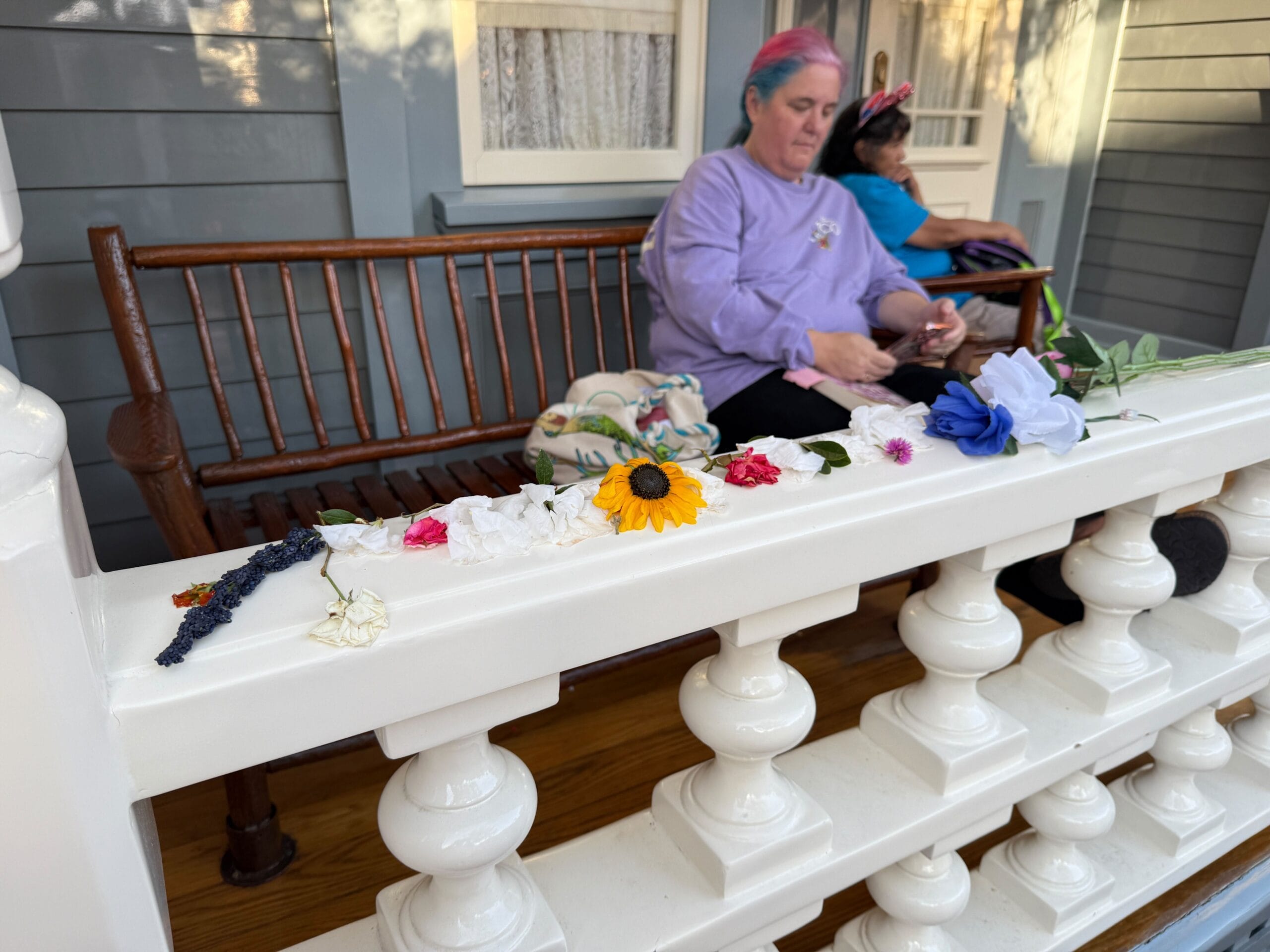 Two people sit on a bench behind a white railing decorated with various colorful flowers, including a yellow sunflower.