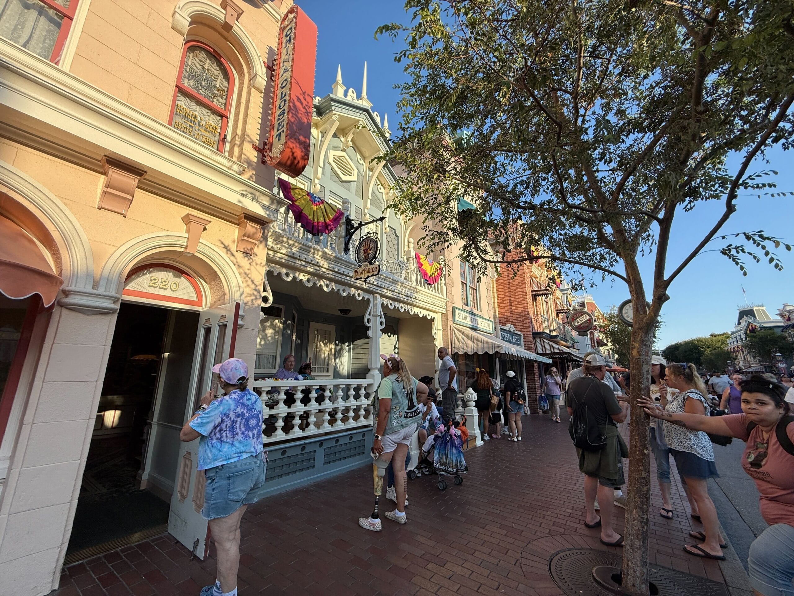 People walk along a brick street lined with shops and trees in a lively theme park setting on a sunny day.