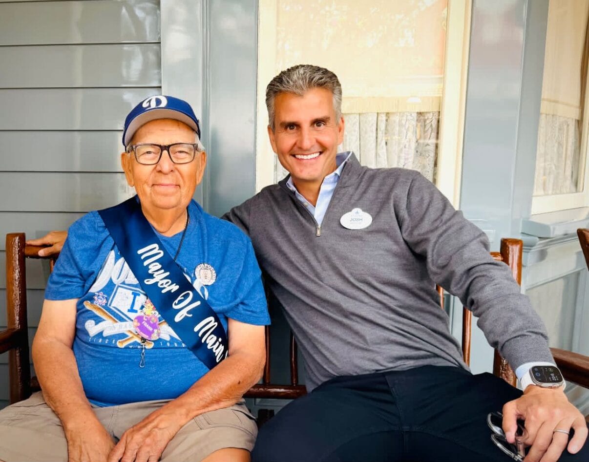Two men sit smiling on a bench at Disneyland; one wears a "Mayor of Main Street" sash, the other has a name tag.