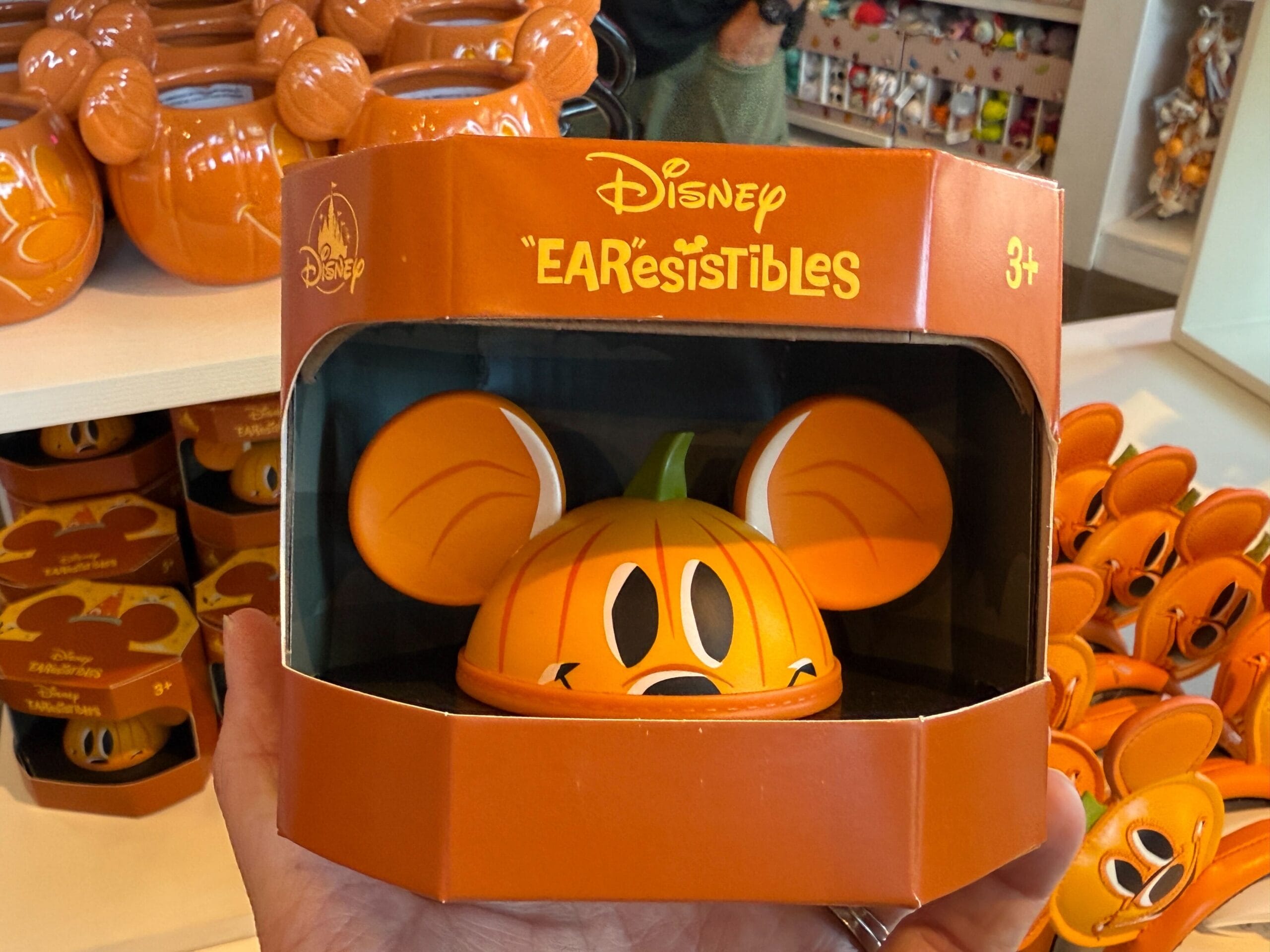 Hand holding a Disneyland “EARresistibles” pumpkin Mickey ear hat, surrounded by Halloween merchandise in orange packaging.