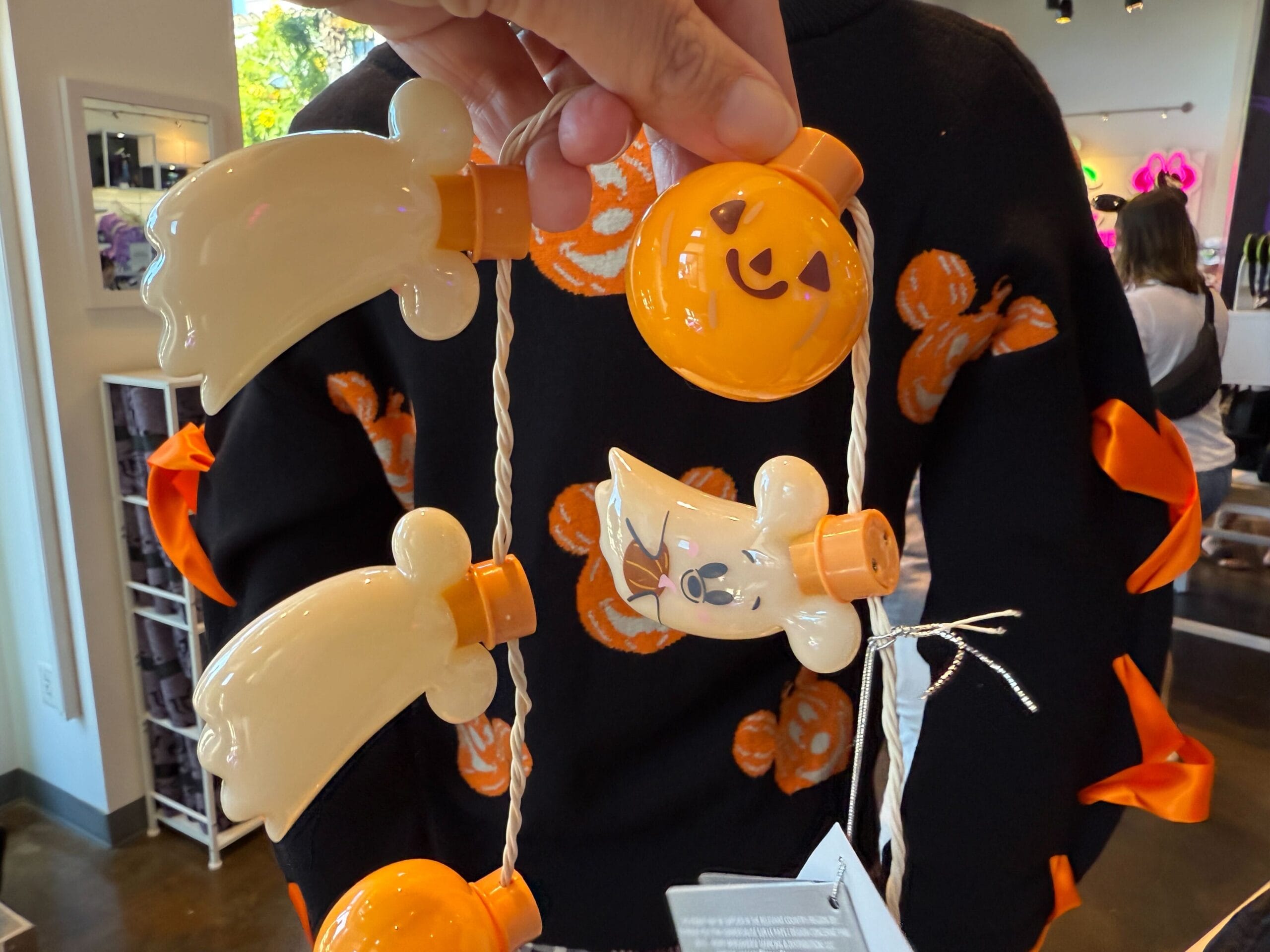 A hand holds Halloween merchandise—a light garland with smiling ghosts and pumpkins—in a brightly lit store.