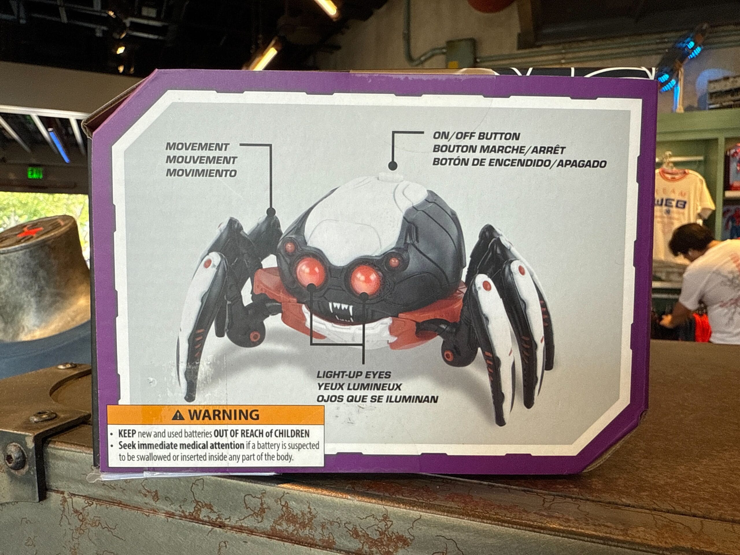 Halloween Spider-Bot toy with red light-up eyes and labeled parts, displayed in packaging with a warning label below.
