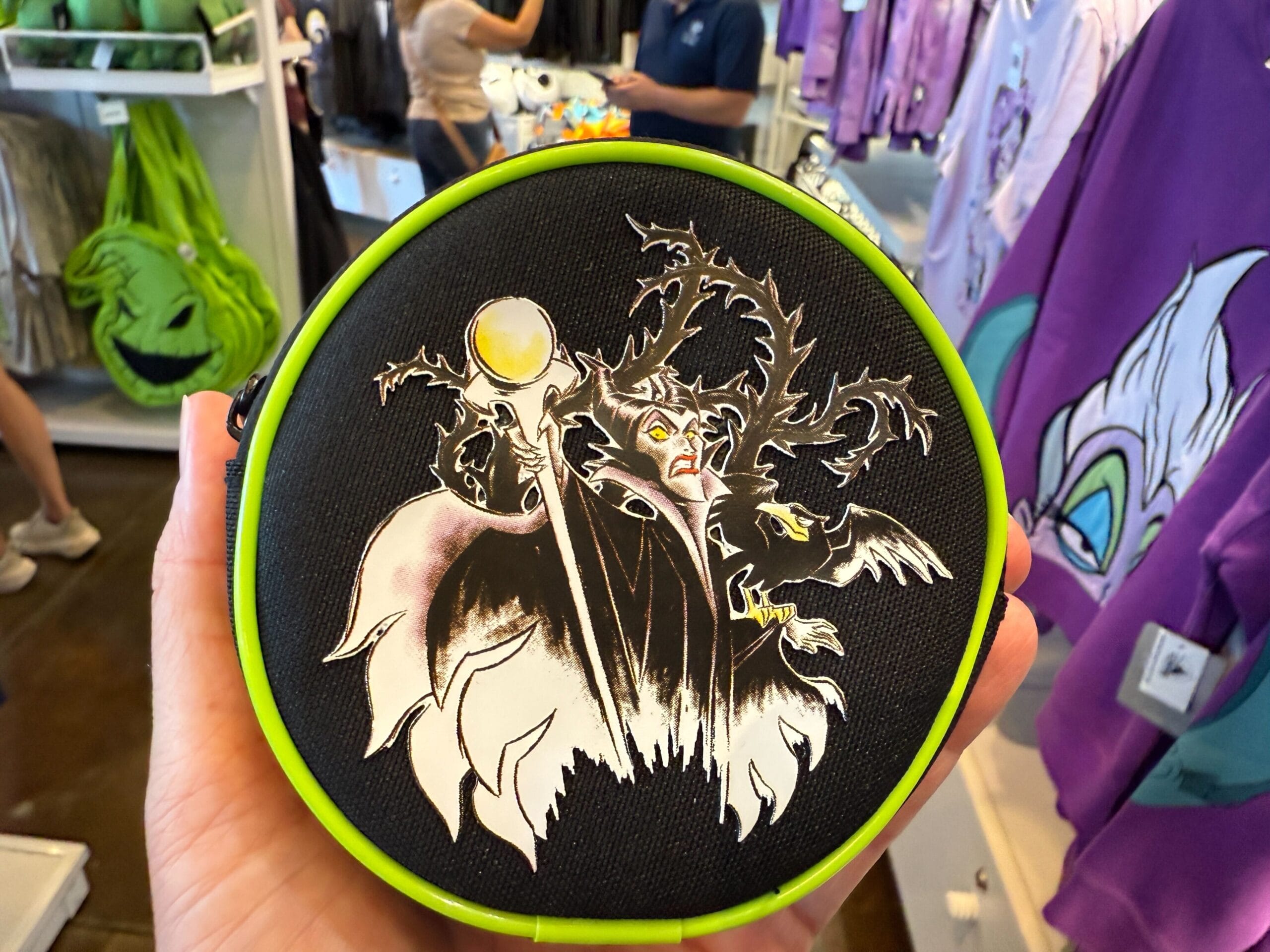 A hand holds Disney Villains apparel—a Maleficent bag with green accents—in a store of villain merchandise.