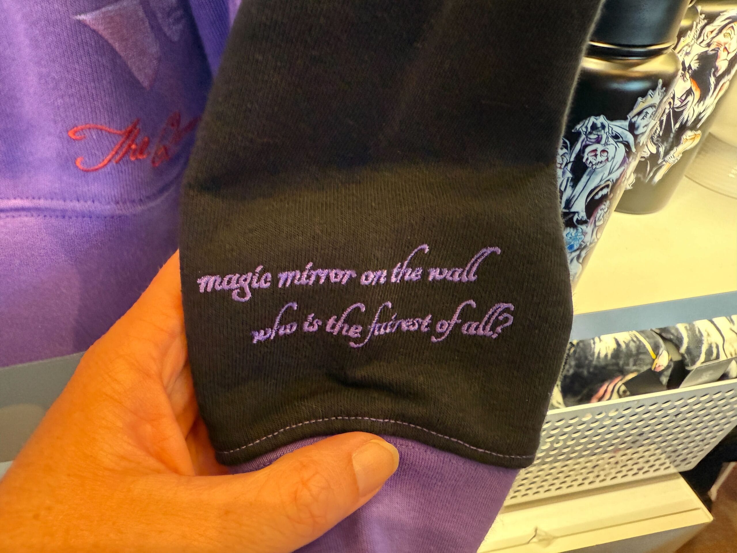 A hand holds Villains Merchandise: a purple and black fabric with "magic mirror on the wall who is the fairest of all?" embroidered.