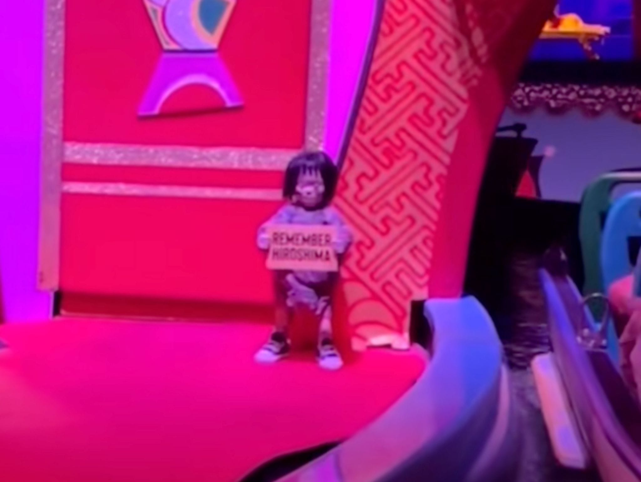 A small doll holds a "Remember Hiroshima" sign in an it's a small world-style red and pink display.