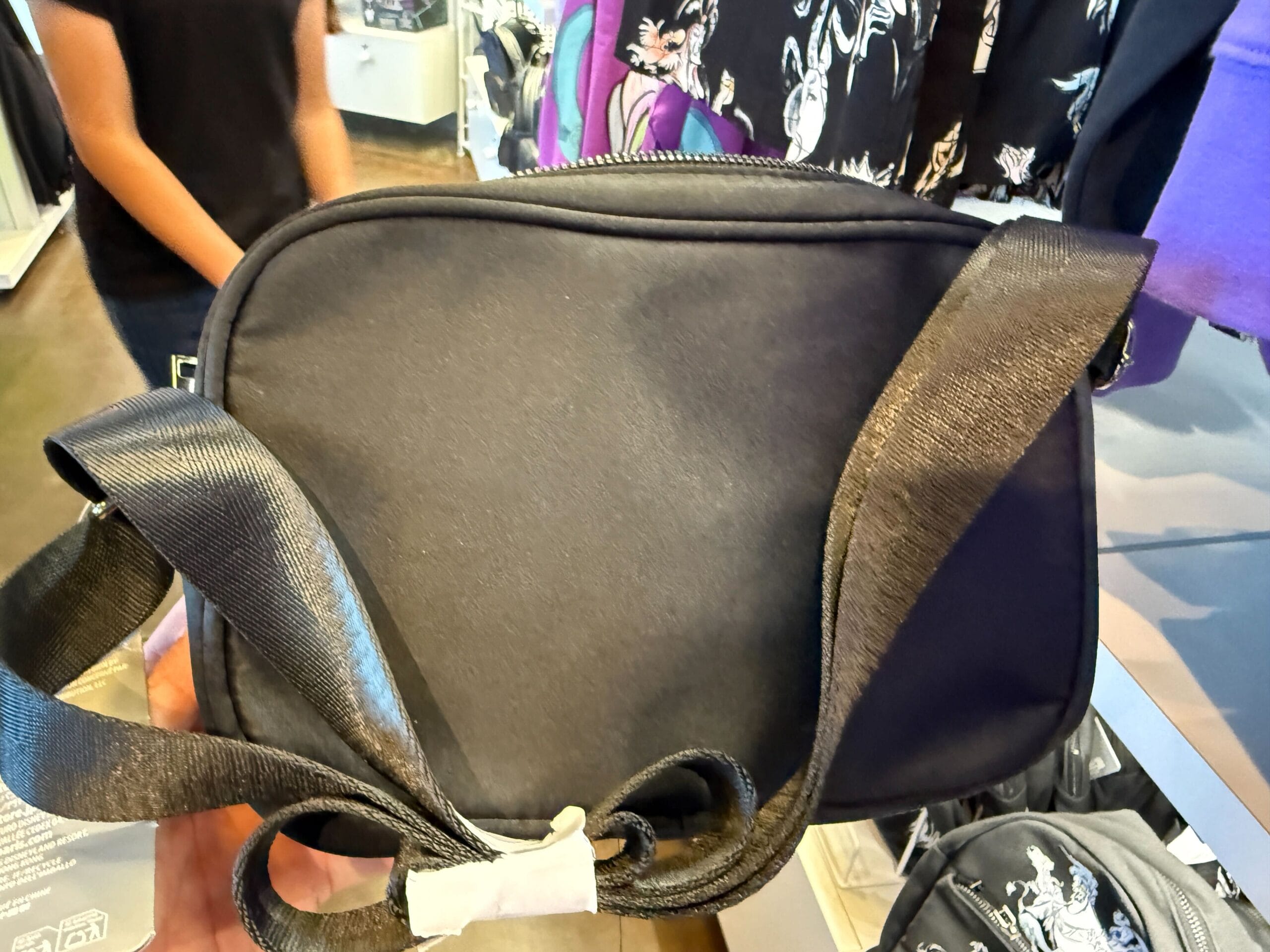 A person holds a small black Villains Apparel shoulder bag with a wide strap in a clothing store.