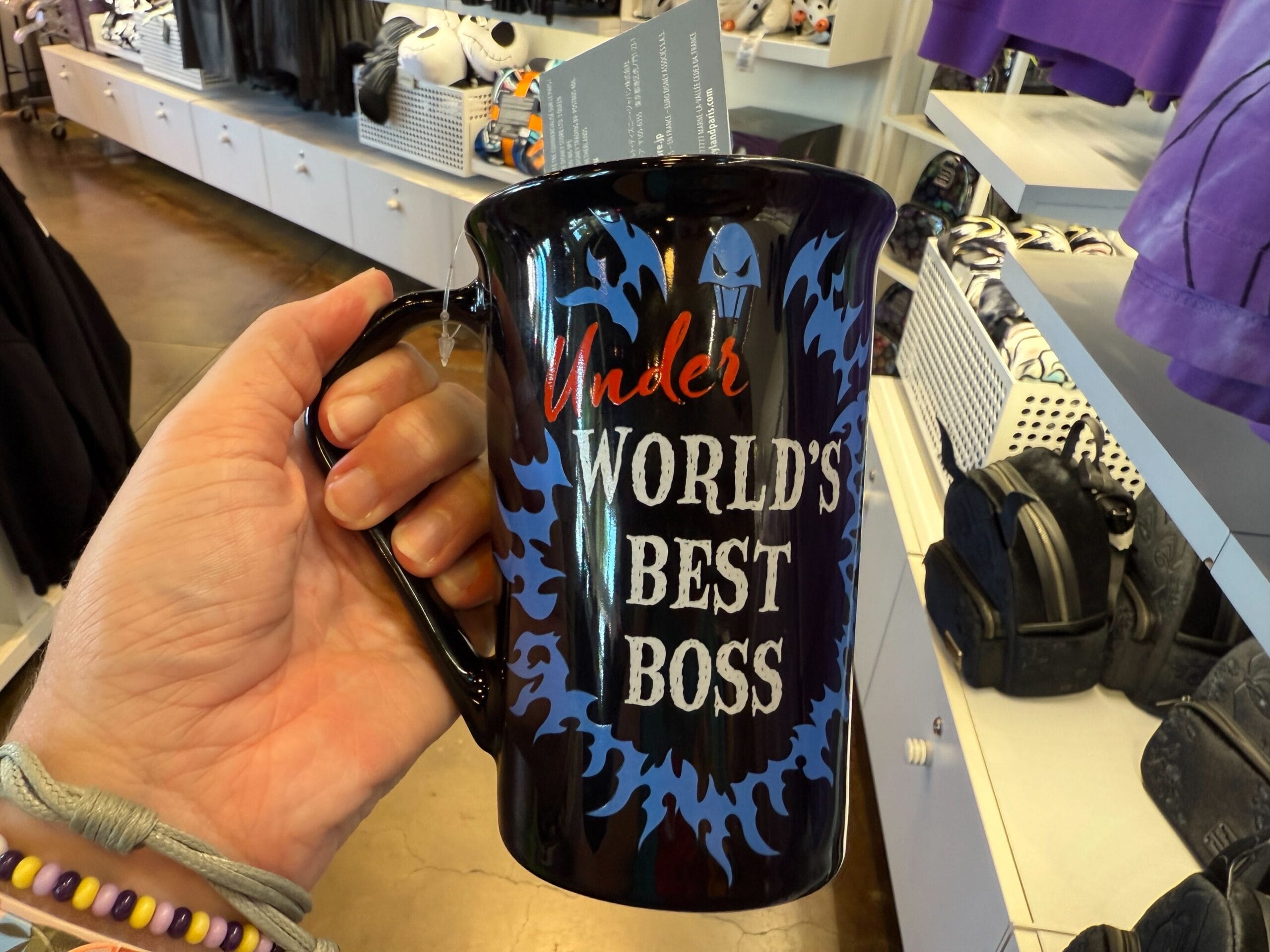 A hand holds a black mug with blue flames labeled "Under WORLD'S BEST BOSS" in Villains Merchandise display.