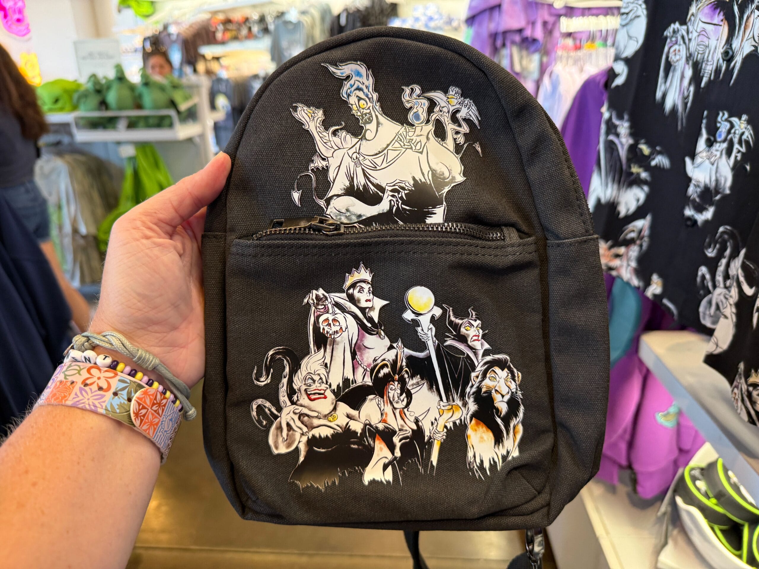 A hand holds Villains Apparel—a black backpack with Disney villains—among colorful merchandise at Disneyland Resort.