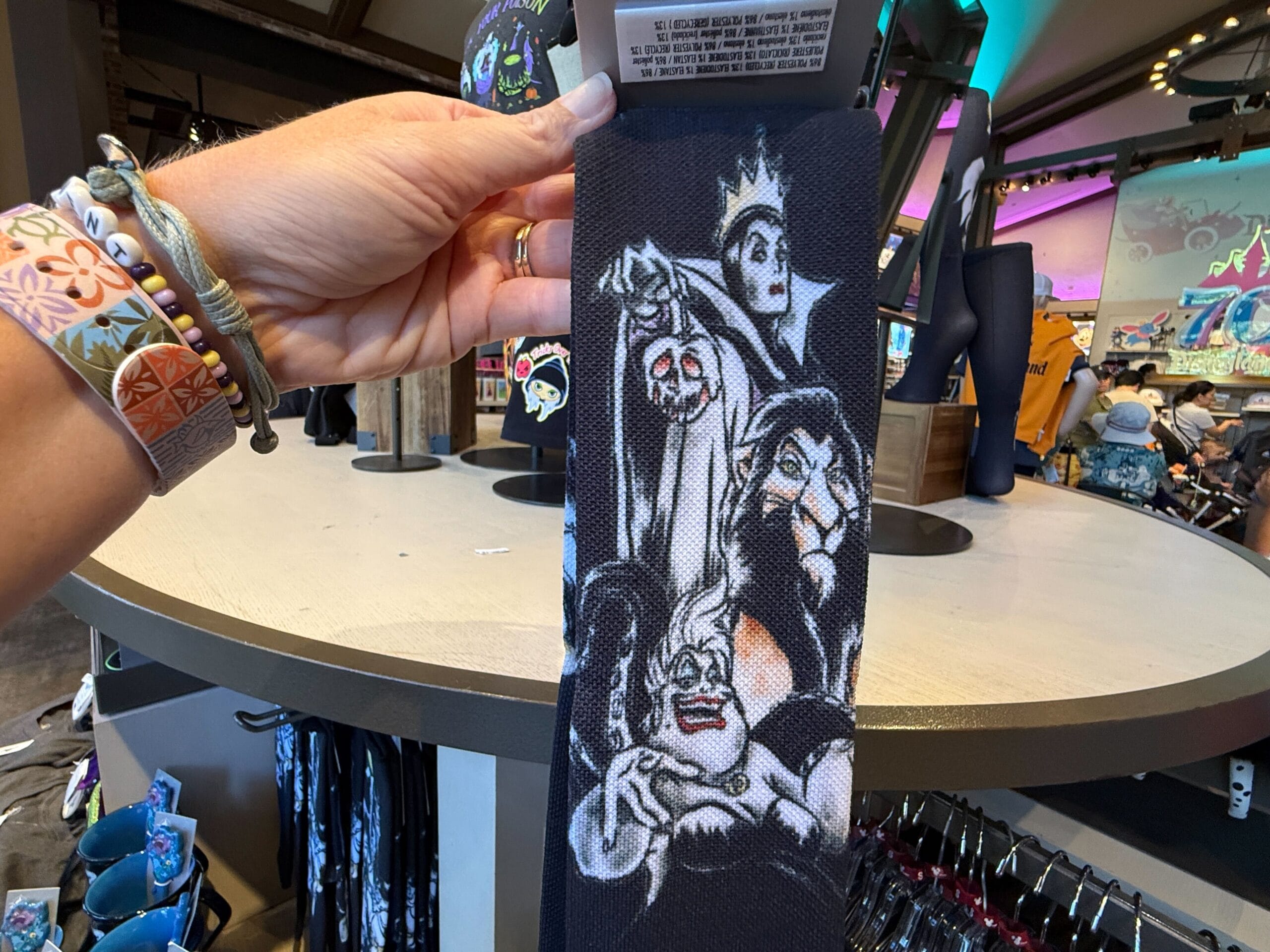 A hand holds up Disney Villains Apparel merchandise featuring Evil Queen, Ursula, and Scar inside a colorful store.