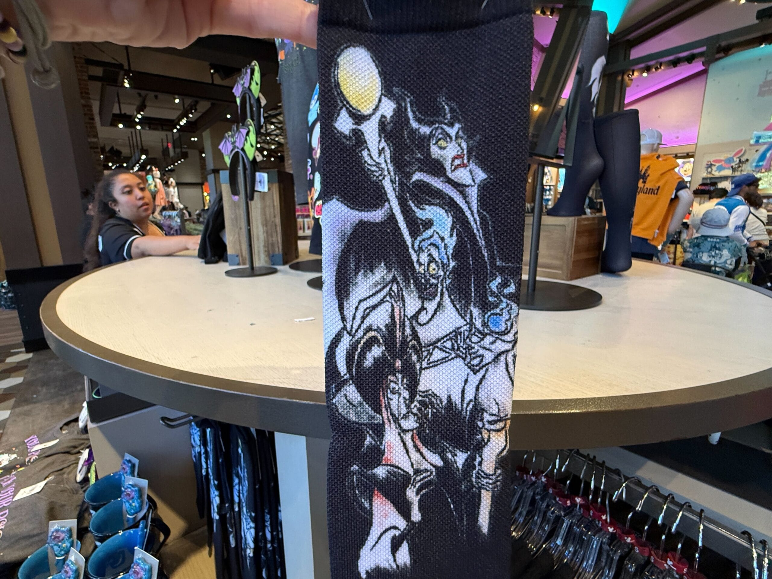 A hand holds Villains Apparel socks with cartoon Disney villains at a Disneyland Resort merchandise store.