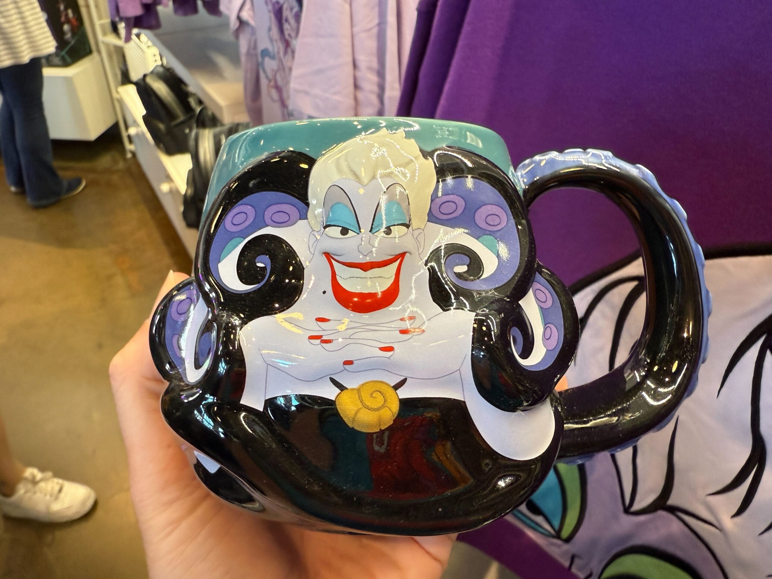 A hand holds a Villains Apparel mug featuring Ursula from The Little Mermaid, with purple tentacles and a big smile.