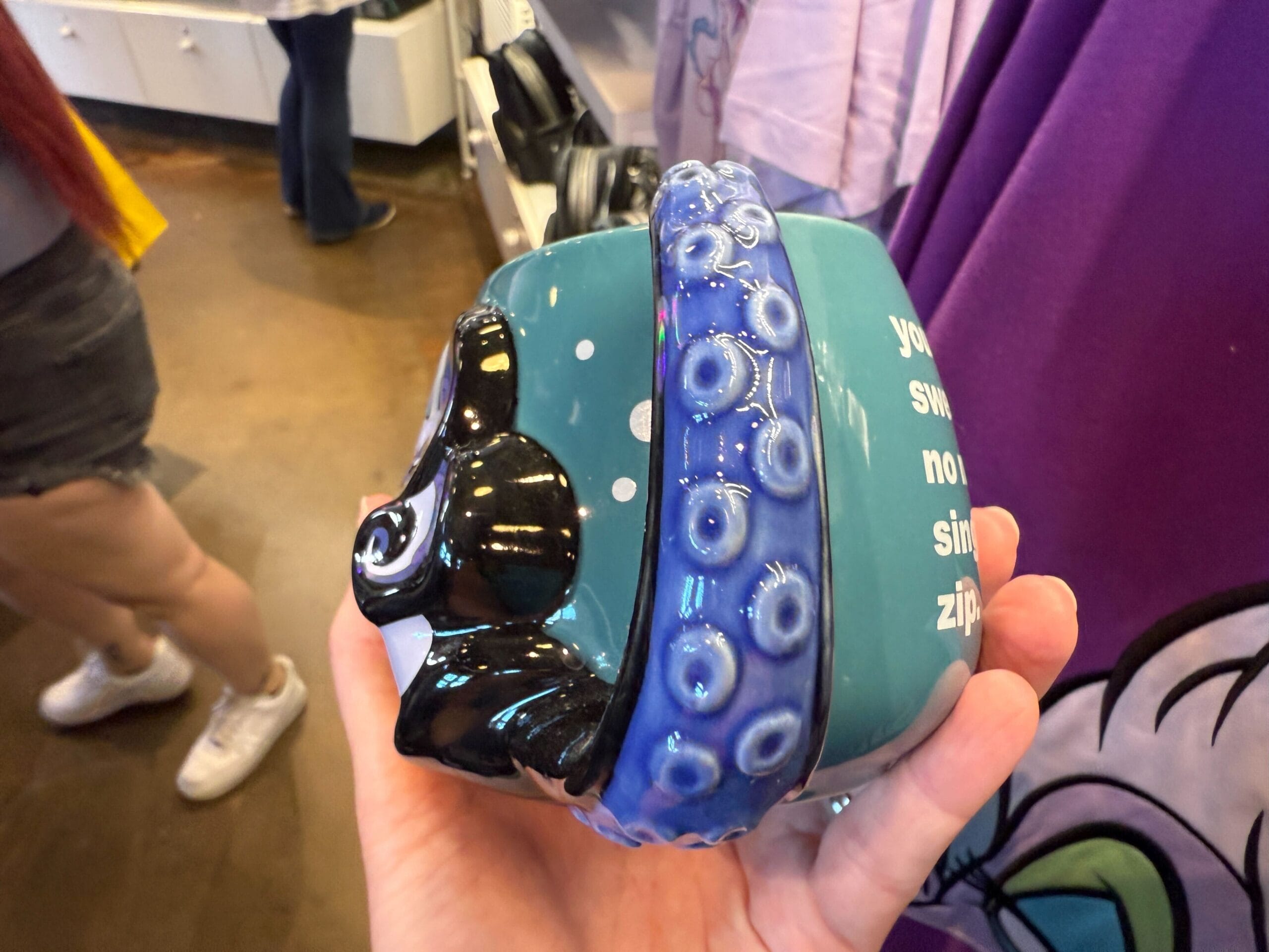 A hand holds a teal Ursula mug from Disneyland Resort, with purple tentacle handle and Villains Merchandise text on the side.