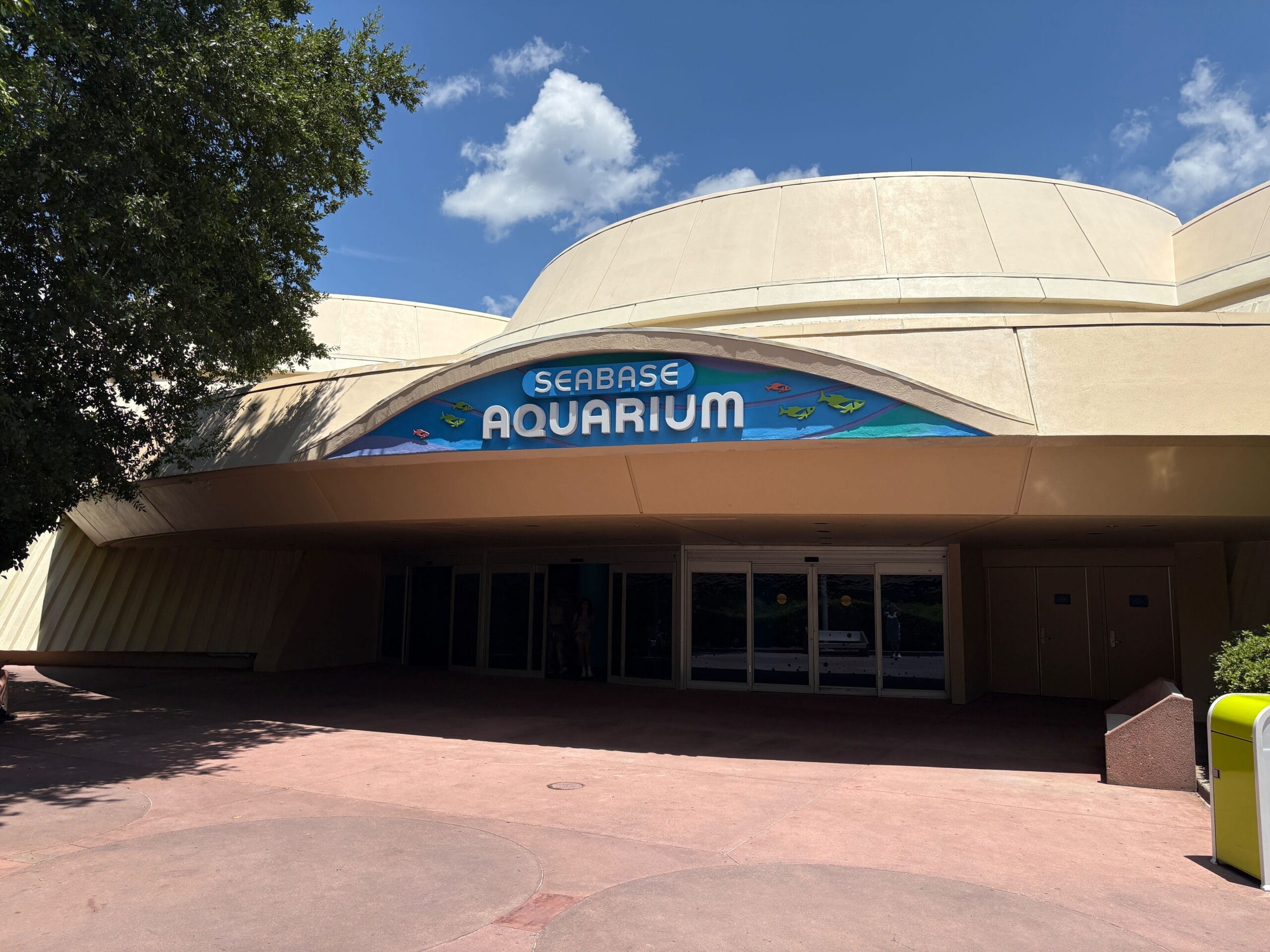The entrance to the Seas Aquarium at EPCOT, with a large sign above glass doors and a tree partially shading the building.