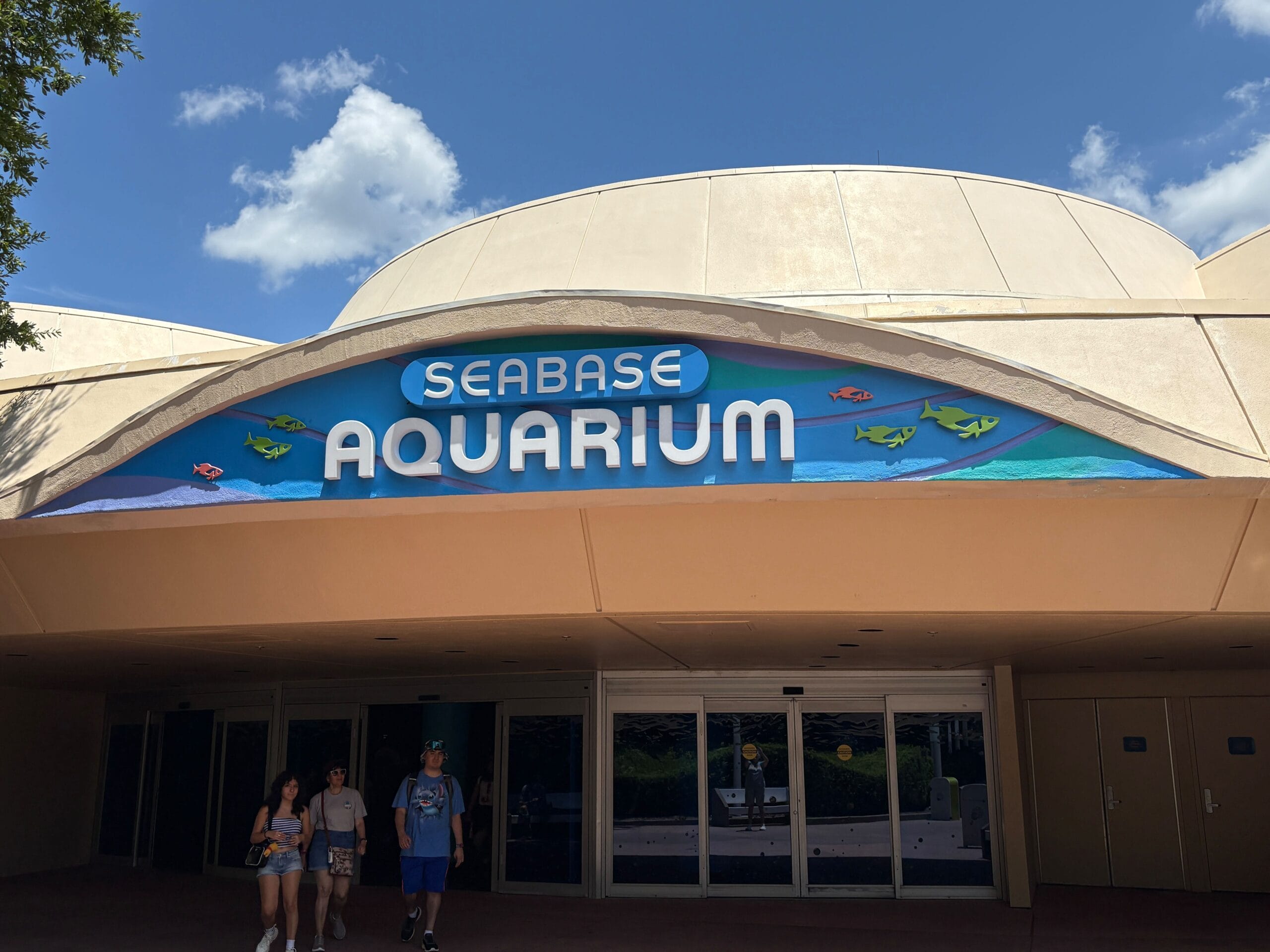 Entrance to Seabase Aquarium at EPCOT features a colorful new aquarium sign under blue skies, as four people walk nearby.