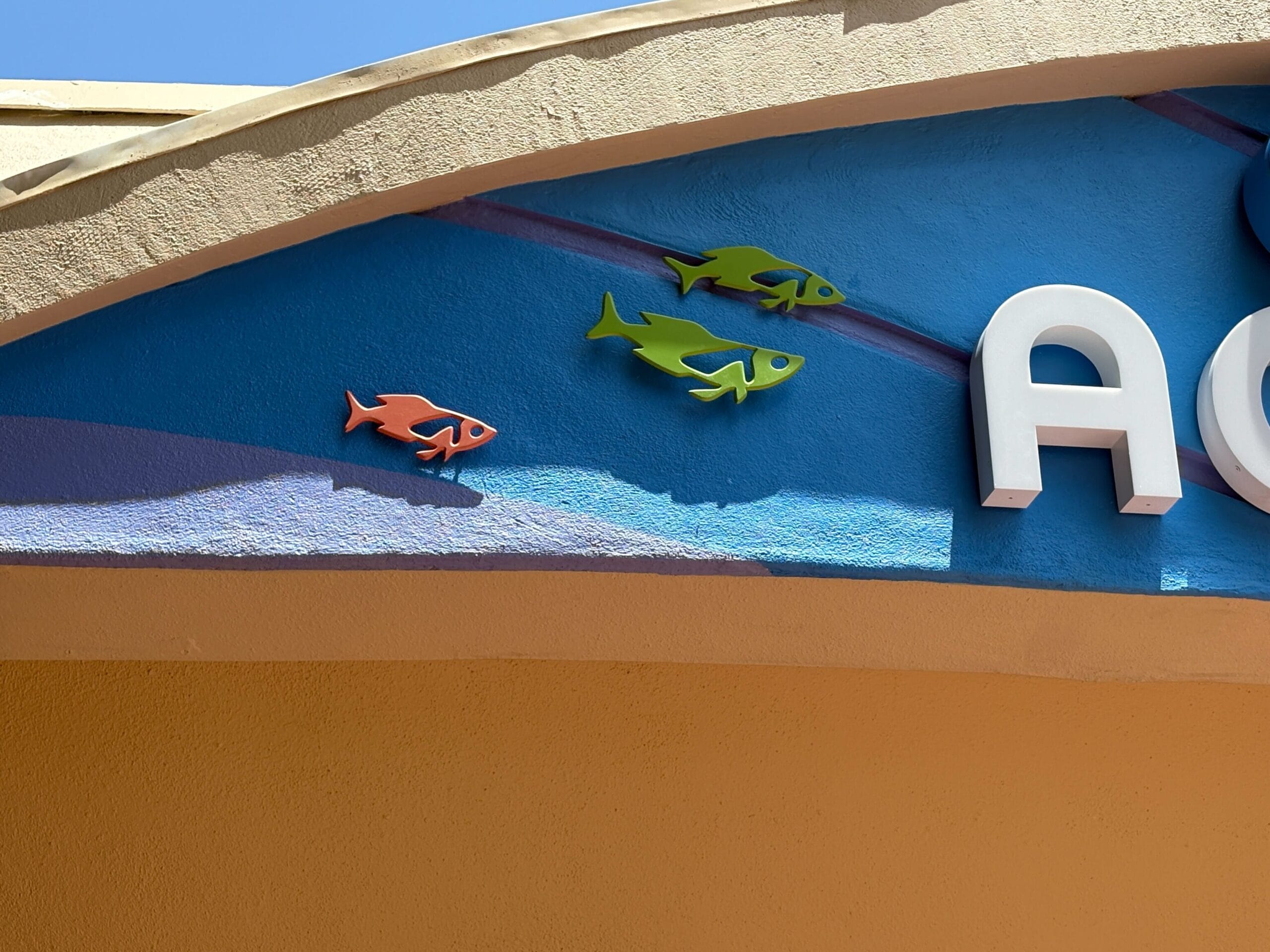 Colorful fish decorations on a blue and yellow wall by the Aquarium Sign, with a clear sky above.
