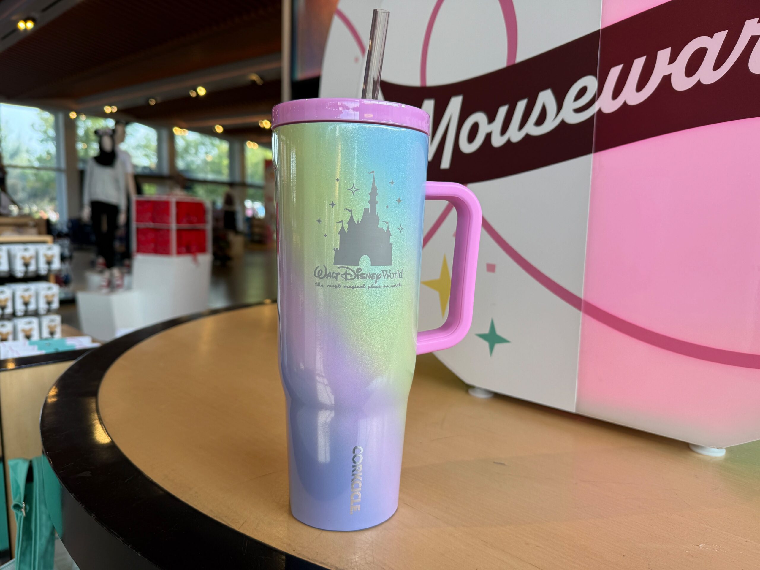 A pastel Disney World travel mug with a handle and straw sits on a counter in a gift shop.