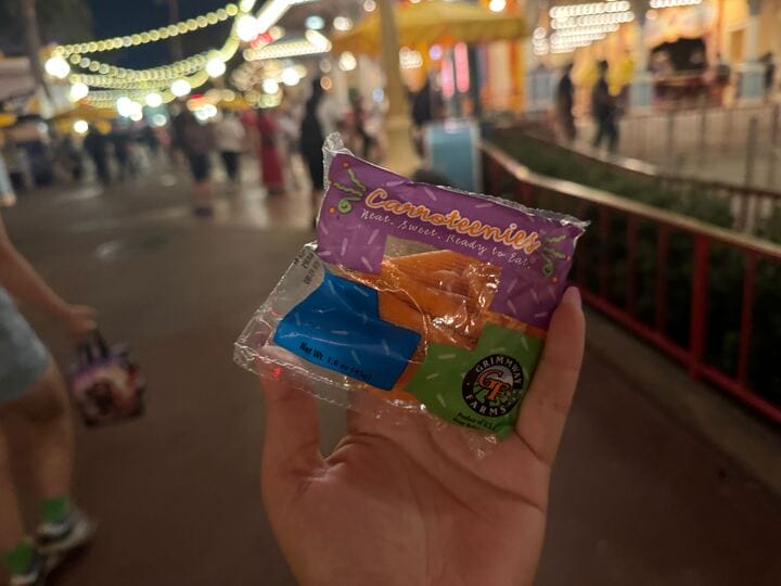 A hand holds a wrapped caramel apple lollipop at night during Oogie Boogie Bash in a lit-up amusement park.