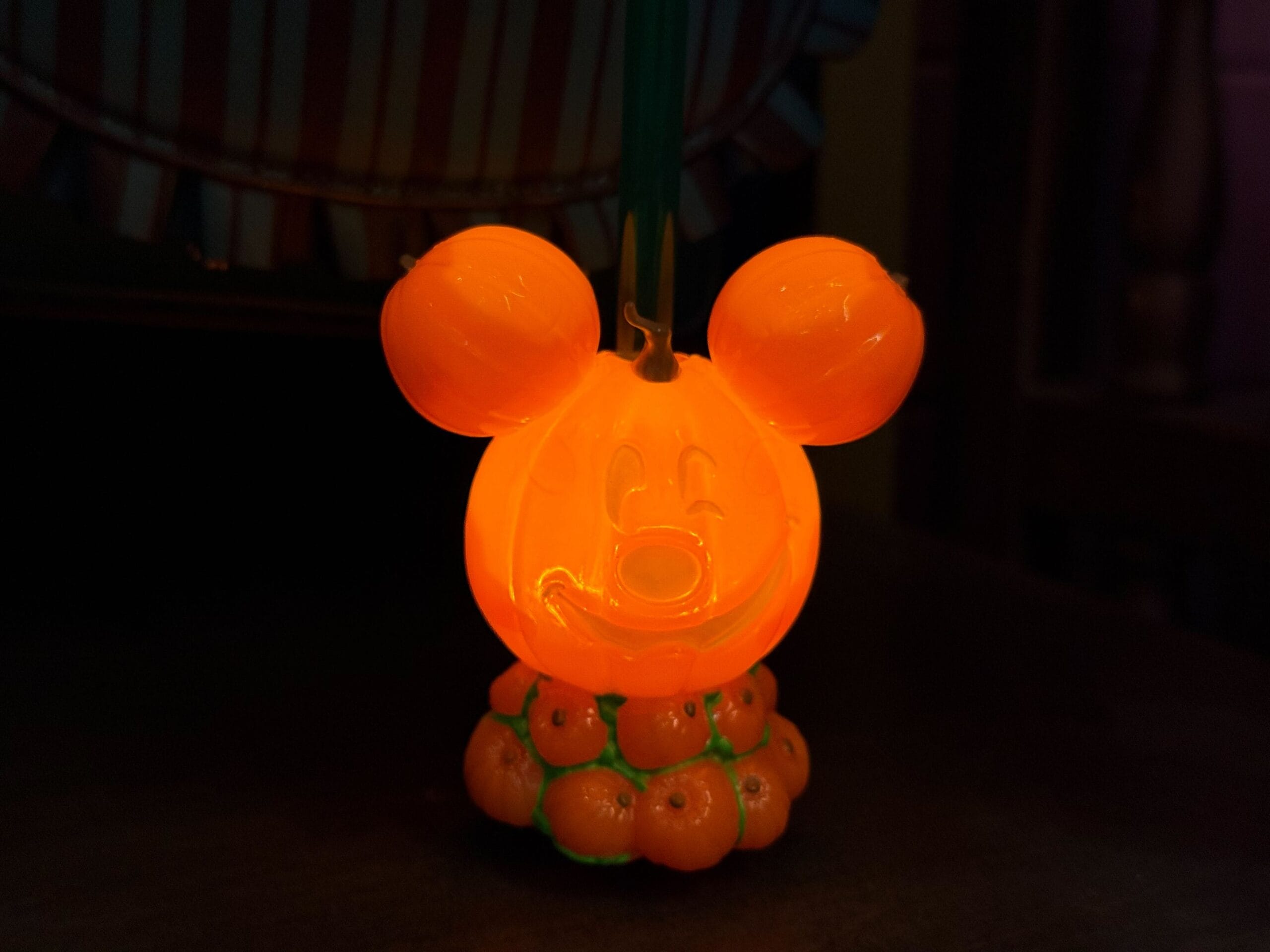 An orange Mickey Mouse-shaped Halloween lantern glows in the dark, sitting on a base of small orange pumpkins.