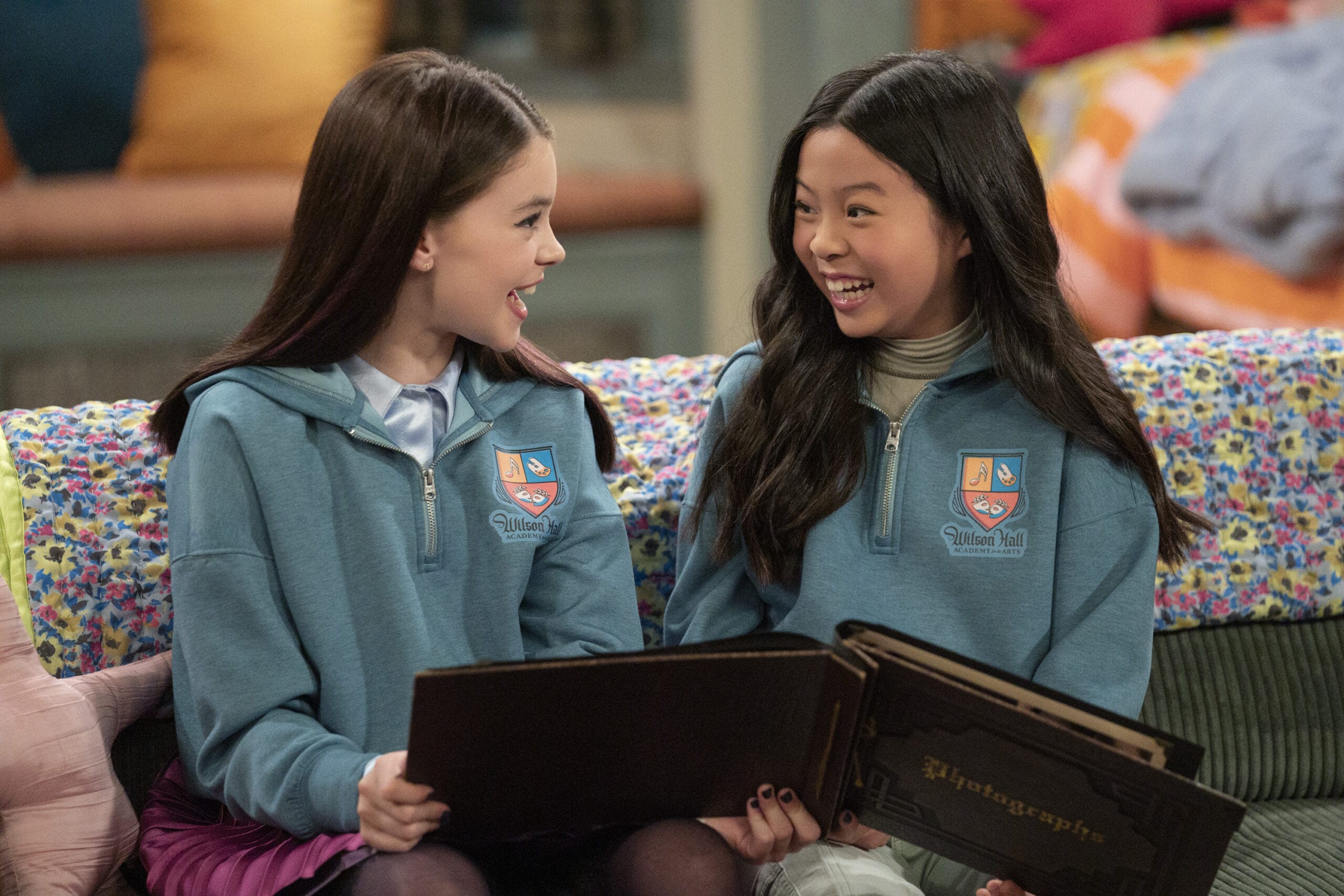 Two girls in matching blue sweaters smile at each other, holding an autograph book from the Live-Action Vampirina series.