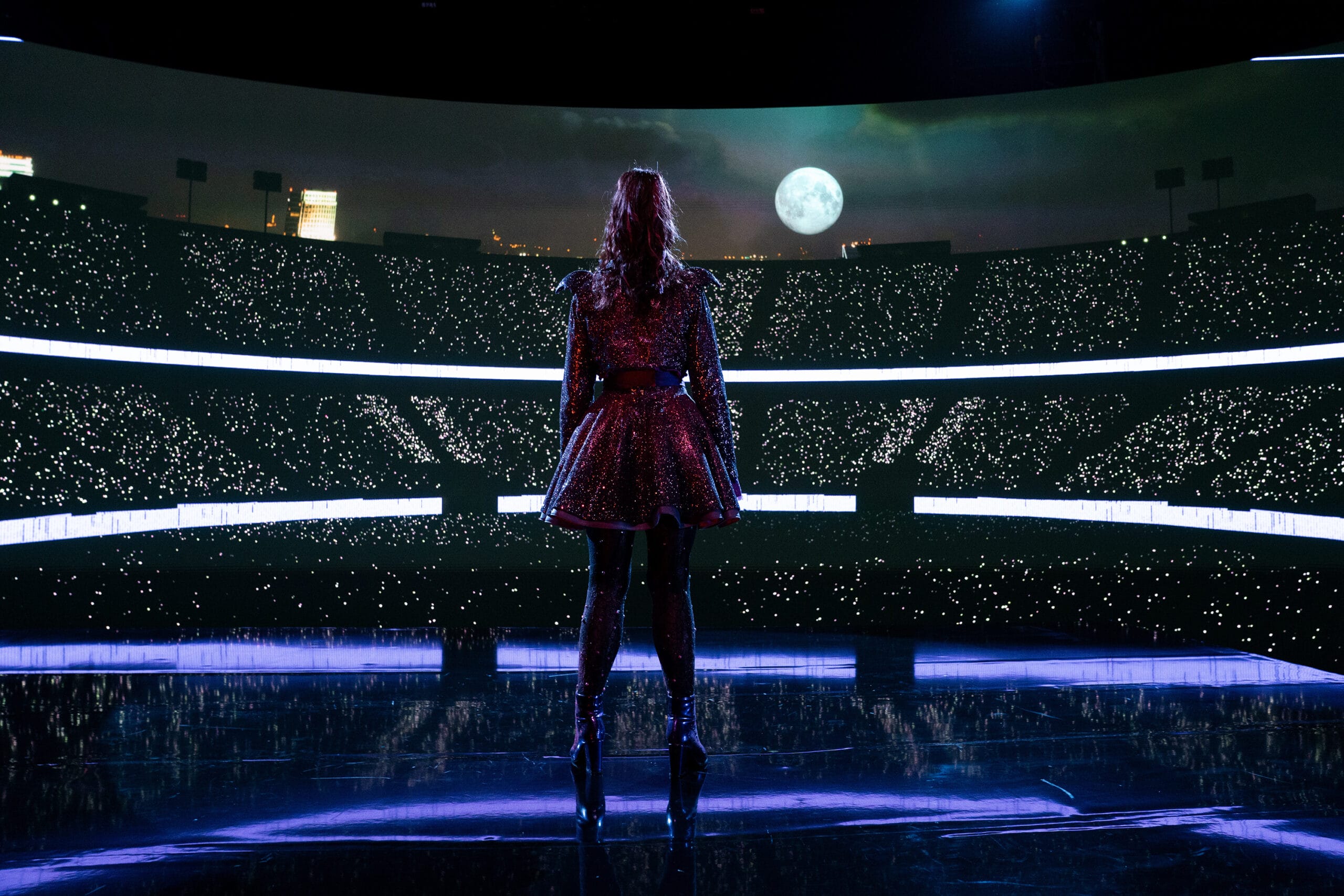 A performer in a sparkly dress stands on stage like Vampirina, facing stadium lights under the moonlit sky.