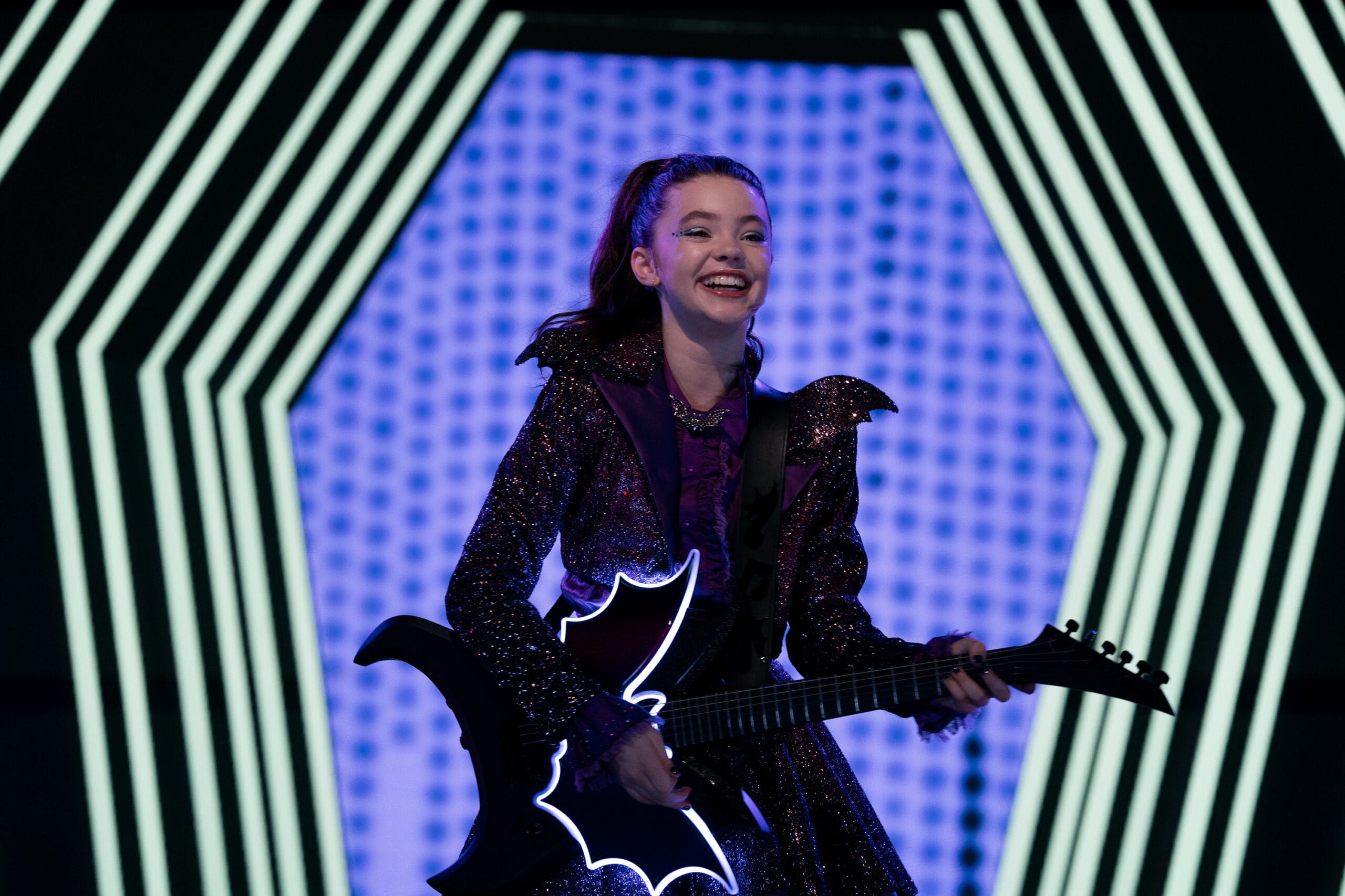 Smiling girl in a sparkly outfit rocks a bat-shaped guitar on a blue-lit stage, like a live-action Vampirina.