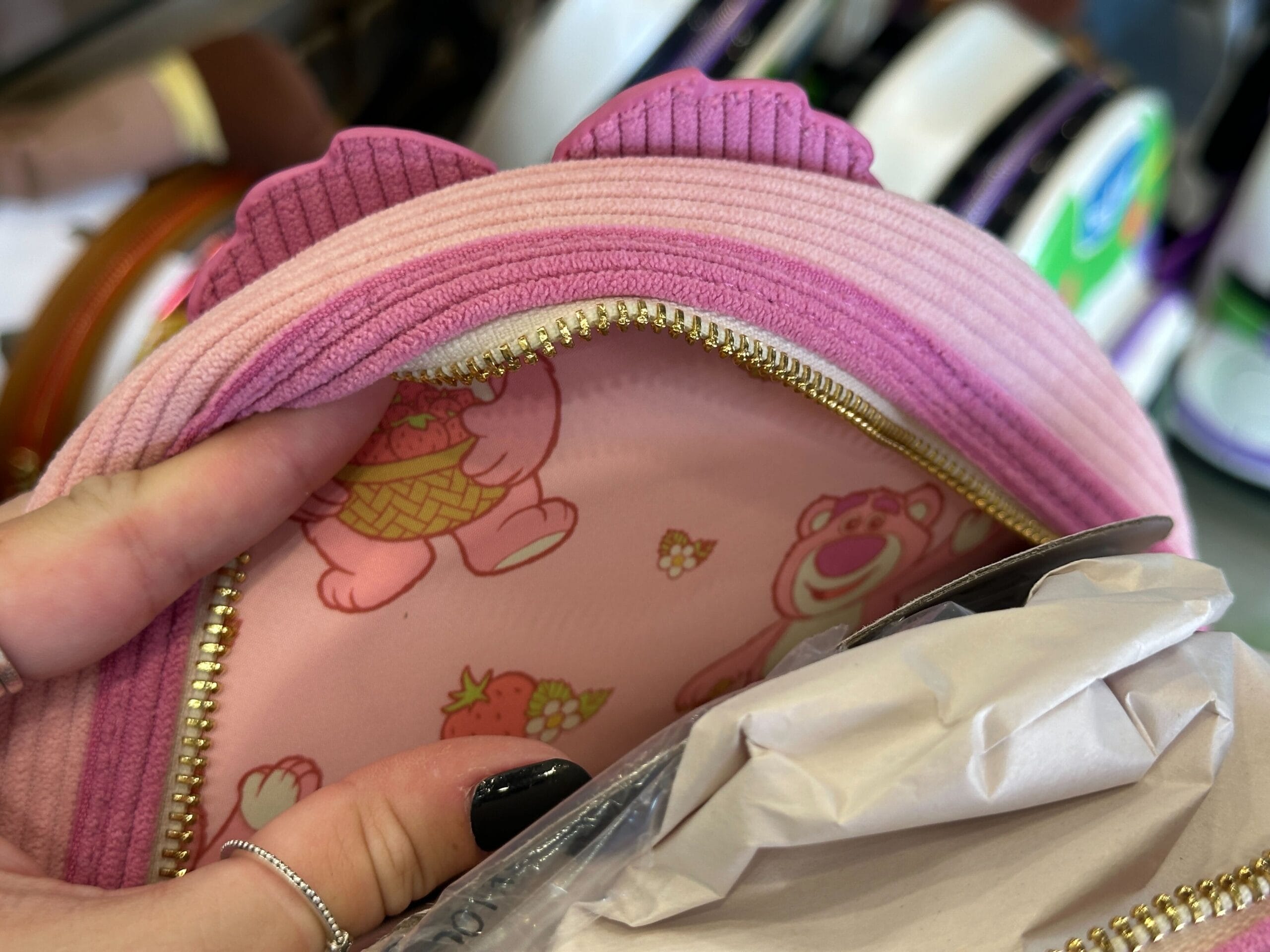 A hand holds a pink Scented Lotso Loungefly Backpack with a bear-print design and purple accents, next to white paper.