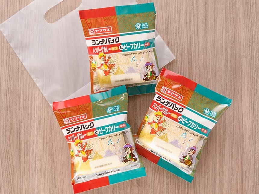 Three packaged Japanese sandwiches, part of a To-Go Lunch Pack, sit on a wooden table near a white plastic bag.