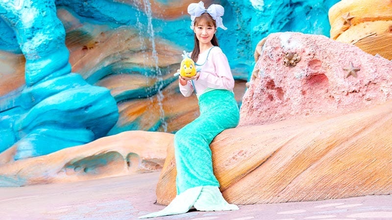 A woman in a mermaid costume sits on a rock with blue and pink backgrounds, holding a yellow fish toy.