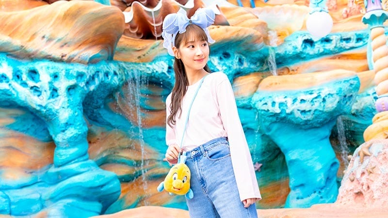 A young woman with blue bow headband and Flounder purse stands in front of a colorful, ocean-themed background.