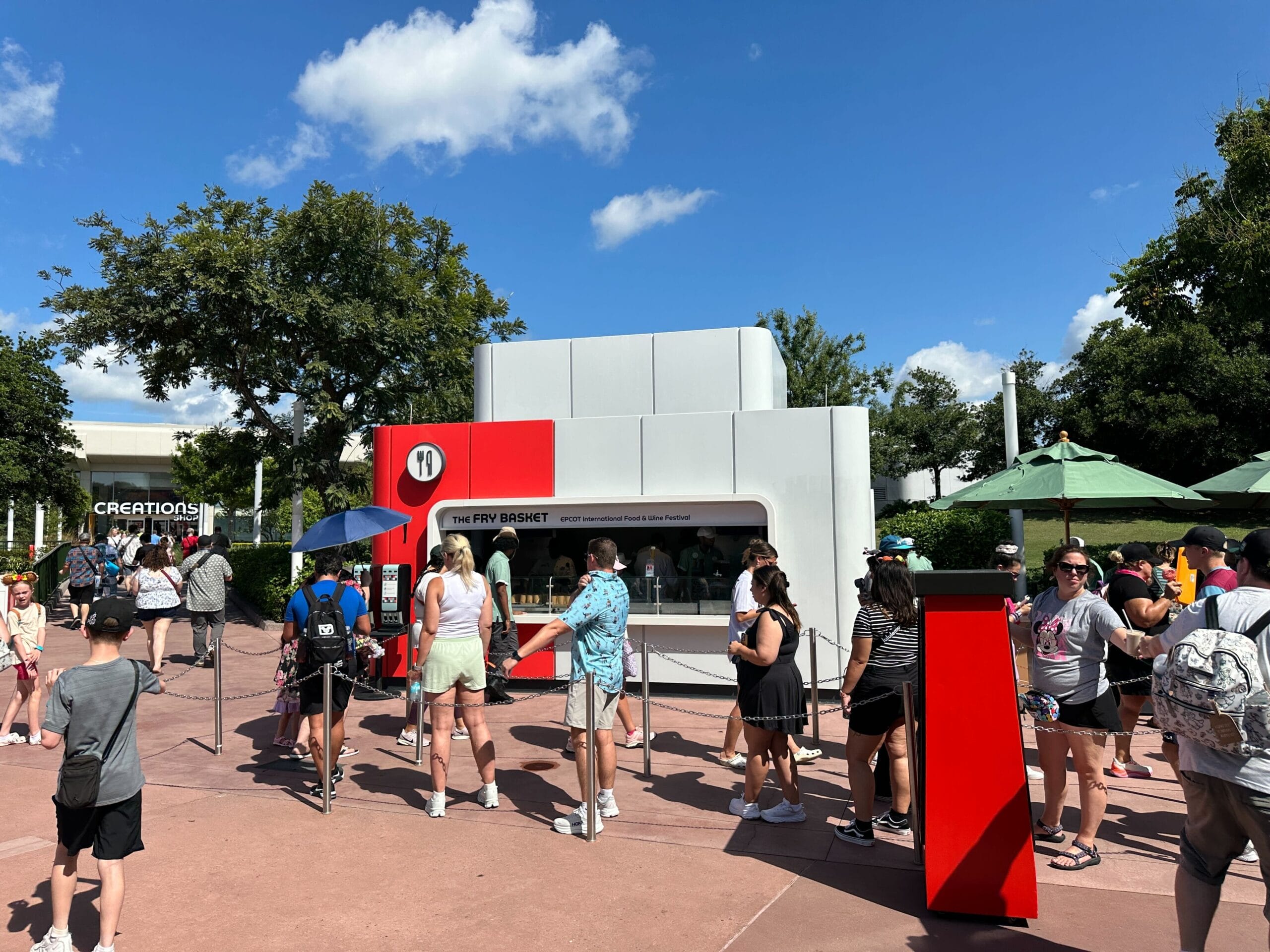People wait in line at The Fry Basket food booth under a blue sky at an outdoor event, with trees and umbrellas nearby.