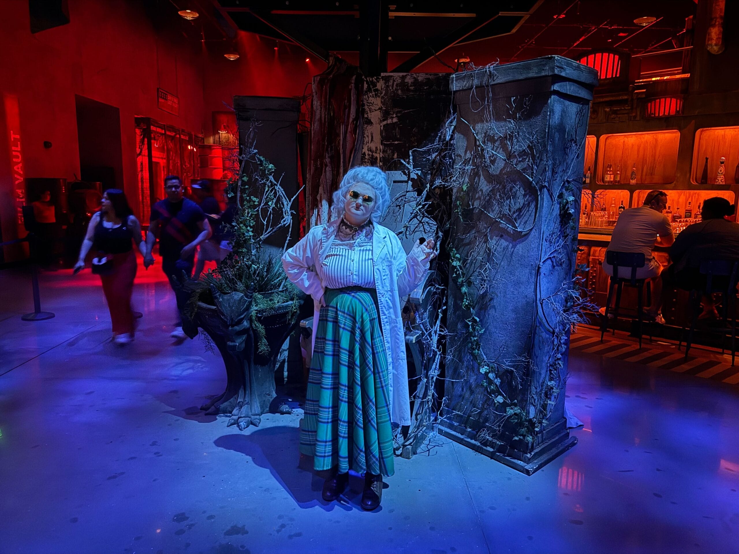 A person in a lab coat and green plaid skirt poses in a spooky, bar-like Las Vegas setting with eerie decorations.