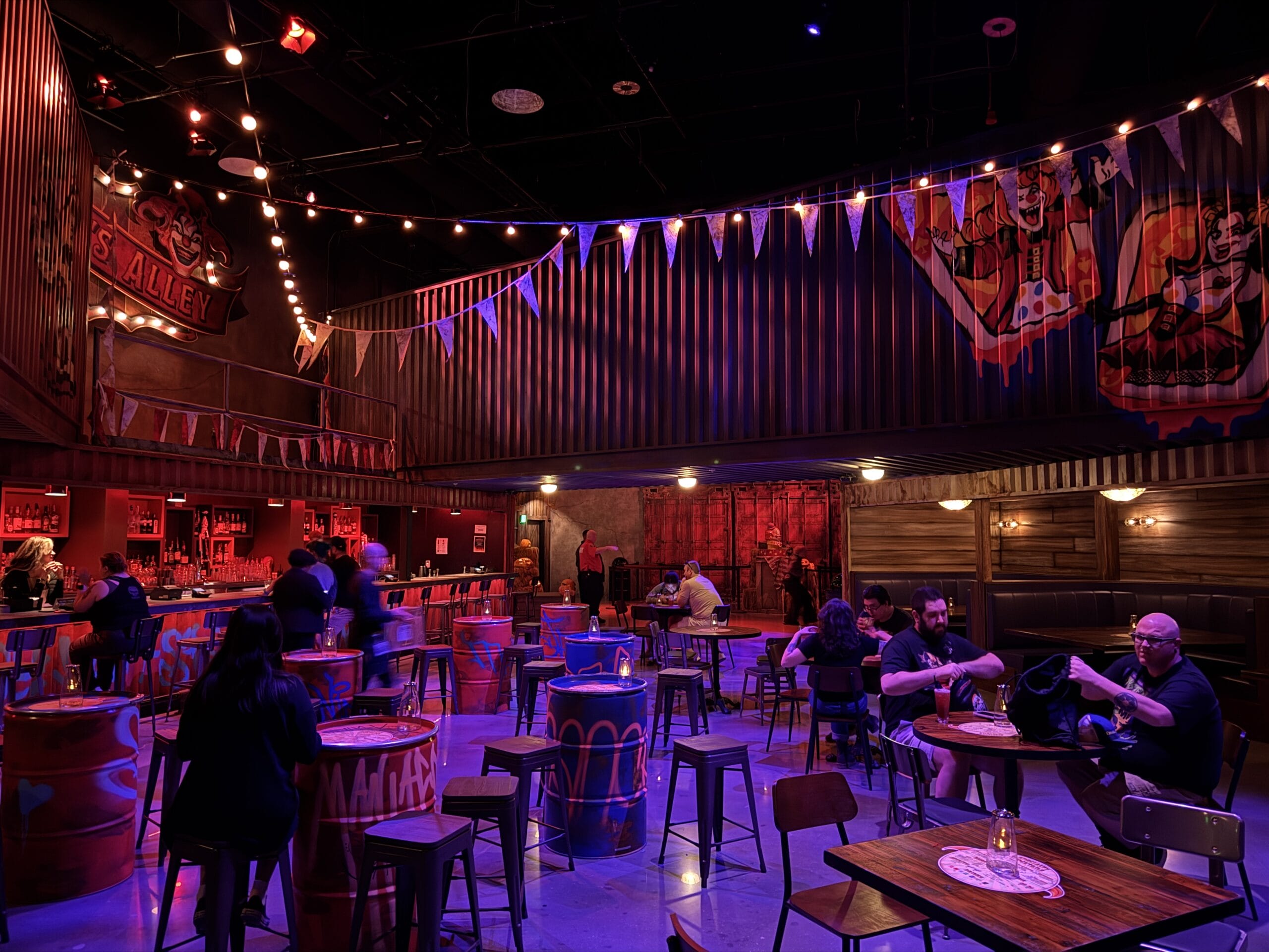 A dimly lit Las Vegas bar with carnival decor, string lights, painted barrels, and people sitting at tables.