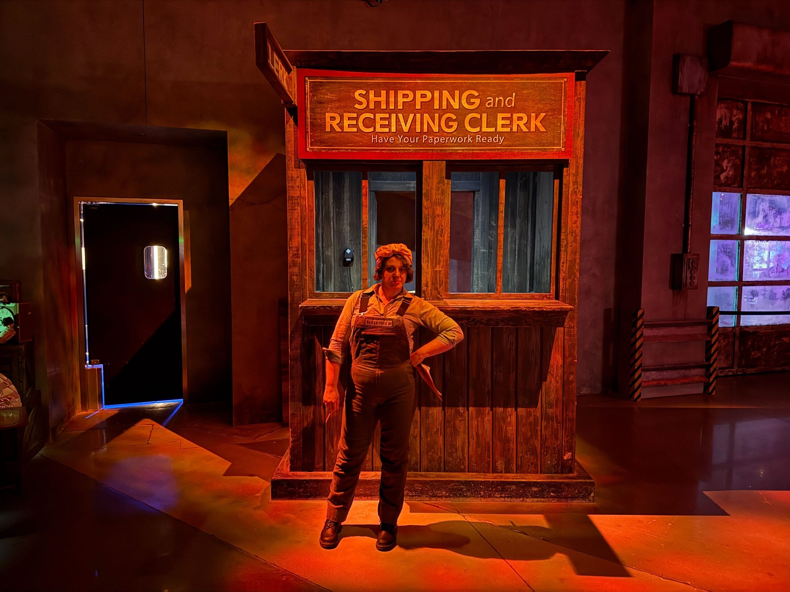Person in overalls stands confidently by a "Shipping and Receiving Clerk" booth at Universal Horror Unleashed, warmly lit.
